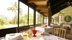 A dining area with tables set for two, large windows with a view of palm trees and mountains, wooden chairs, and a ceiling fan. - Castle at Princeville (Princeville)