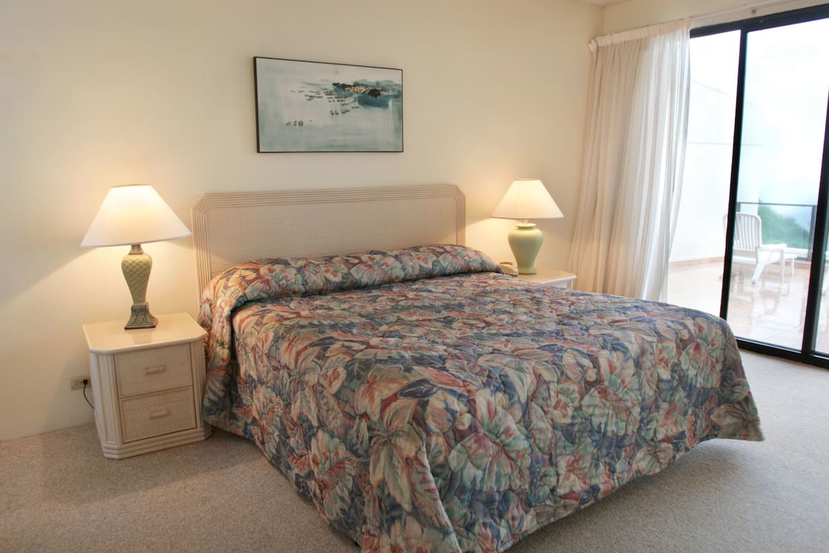 A bedroom with a bed, two bedside tables with lamps, a painting on the wall, and a view through a window.