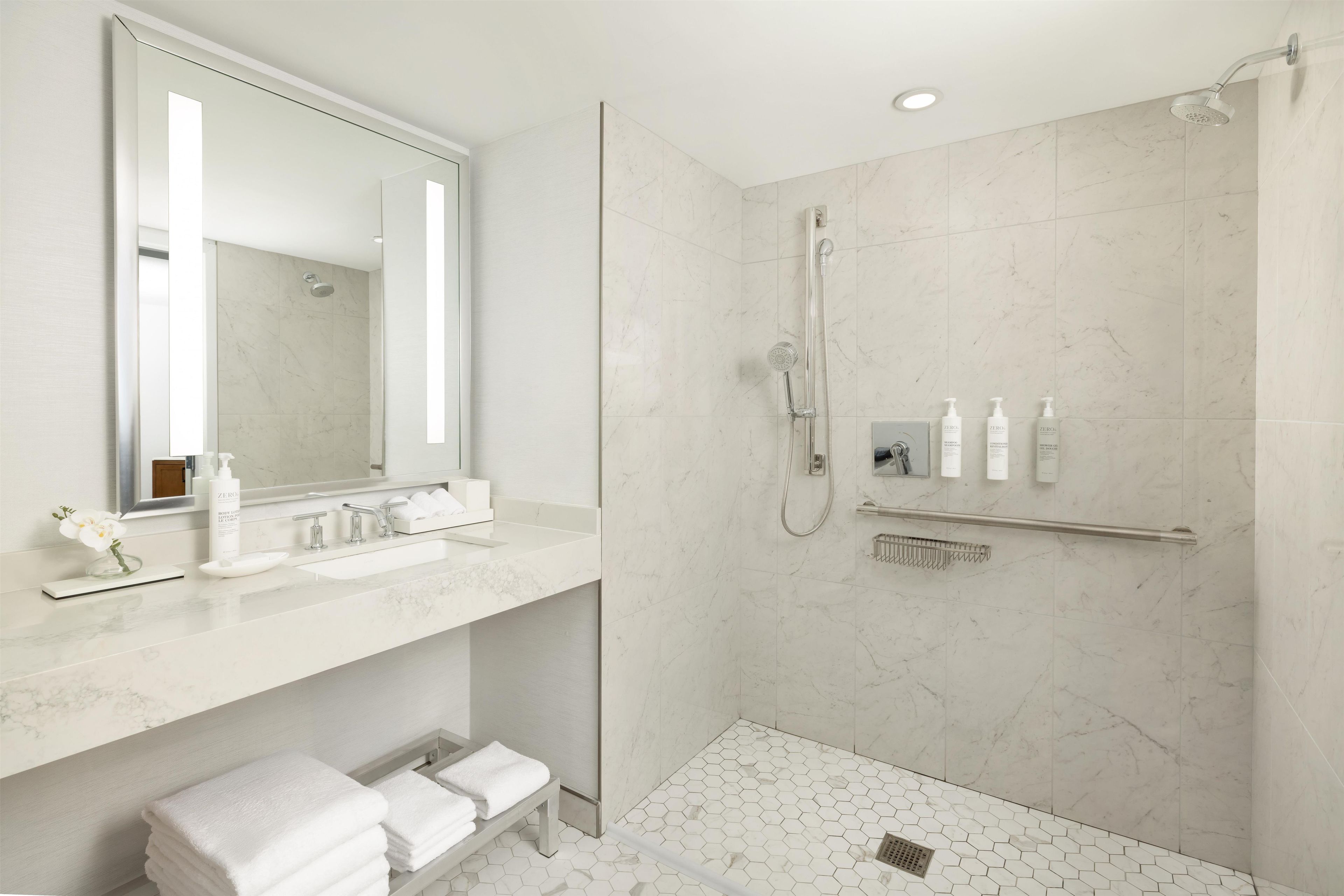 Un baño moderno con un espejo grande, ducha con cabezal de ducha manual y tocador con lavamanos y perchero.