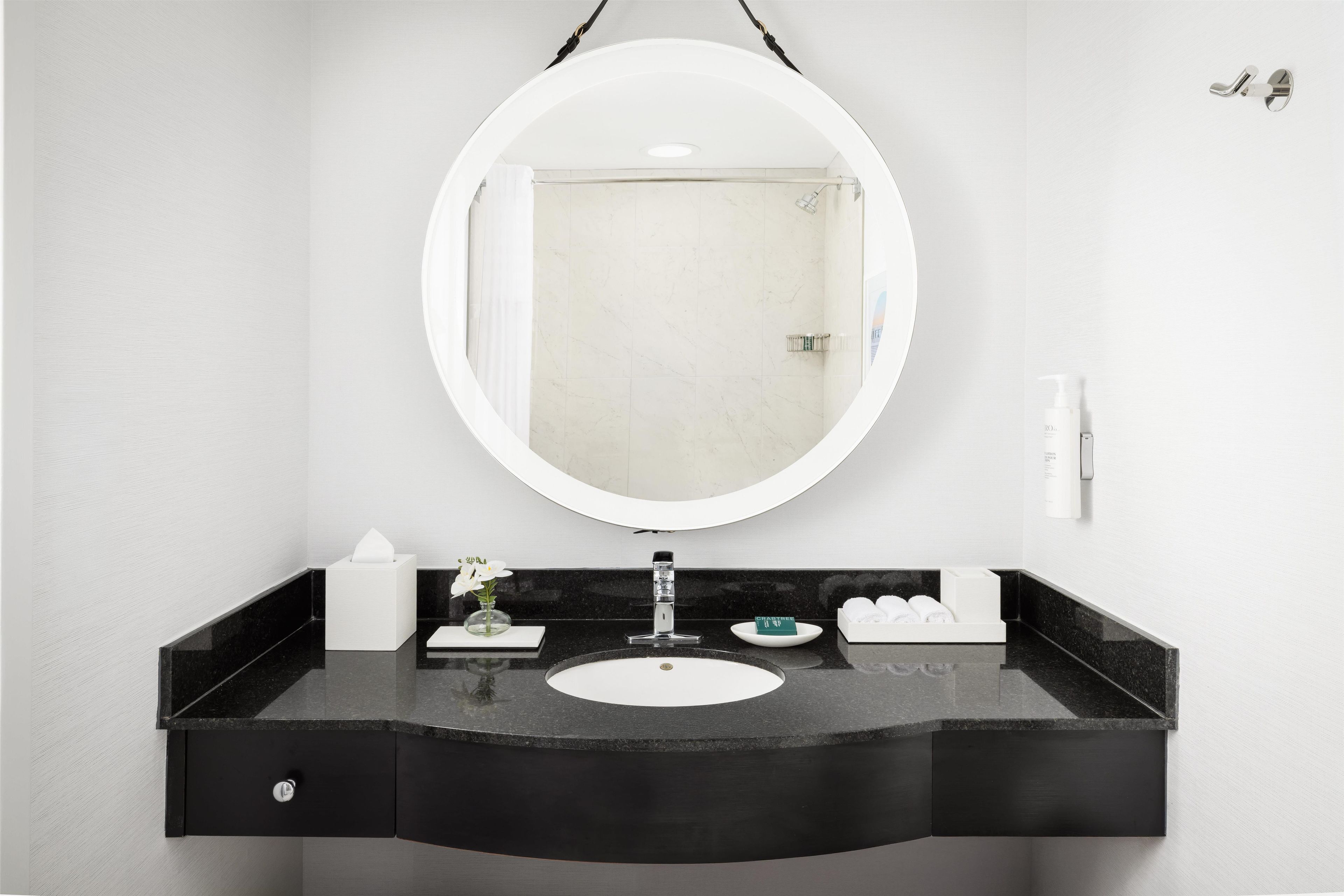 Un baño con tocador negro, espejo redondo, lavamanos blanco y ducha visible a través del espejo.