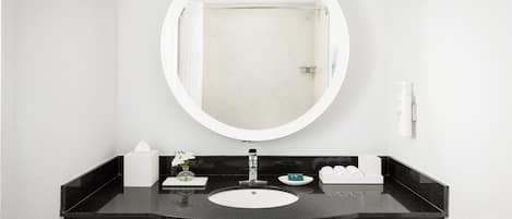 Un baño con tocador negro, espejo redondo, lavamanos blanco y ducha visible a través del espejo.