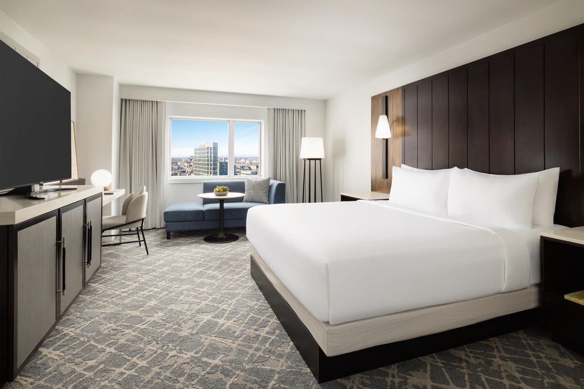 Een hotelkamer met een groot bed, een bureau met een televisie, een kleine tafel met een lamp, een bank en uitzicht op de stad.