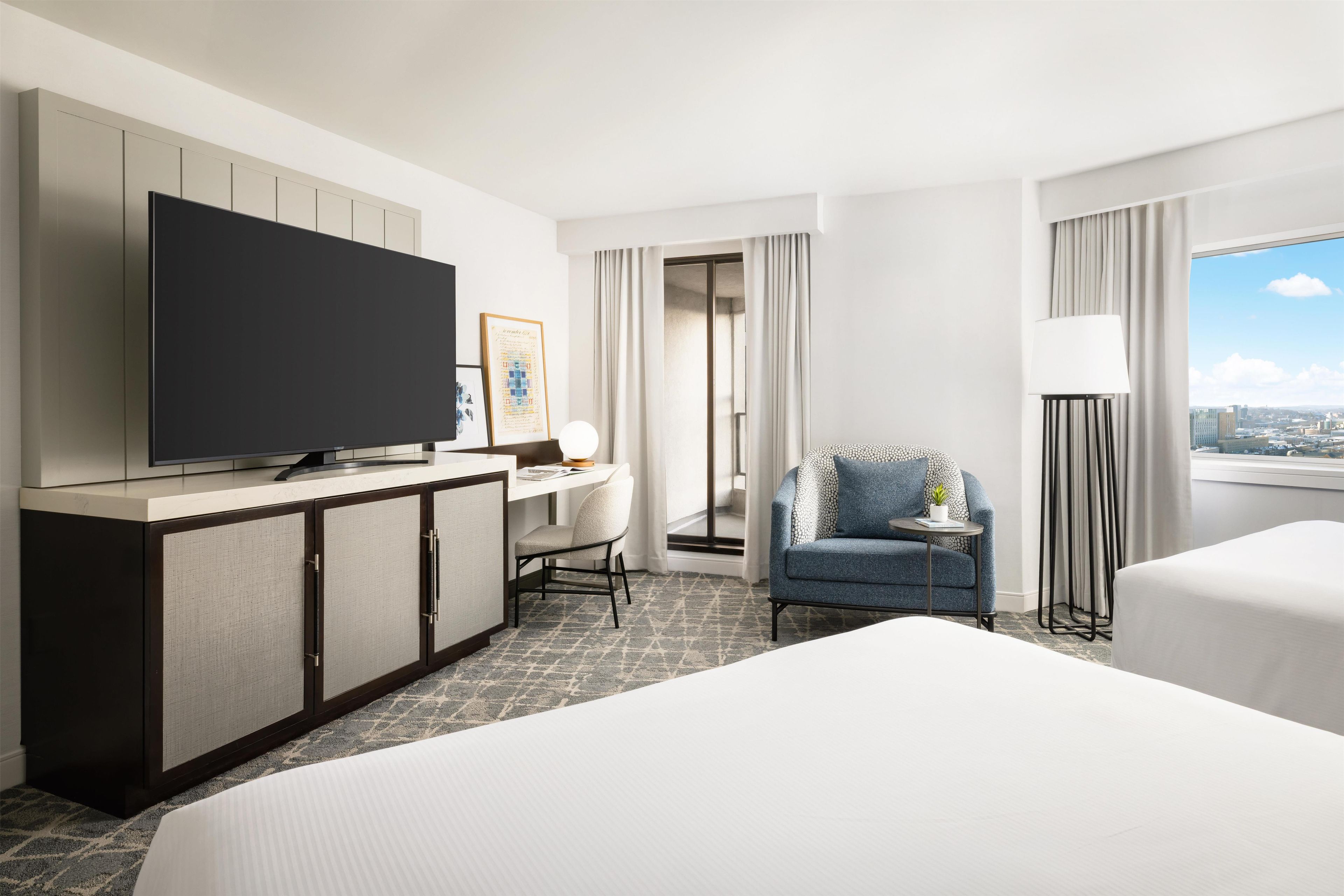 A hotel room with a bed, a desk with a chair, a television, a small table with a lamp, and a view of the city through the window.