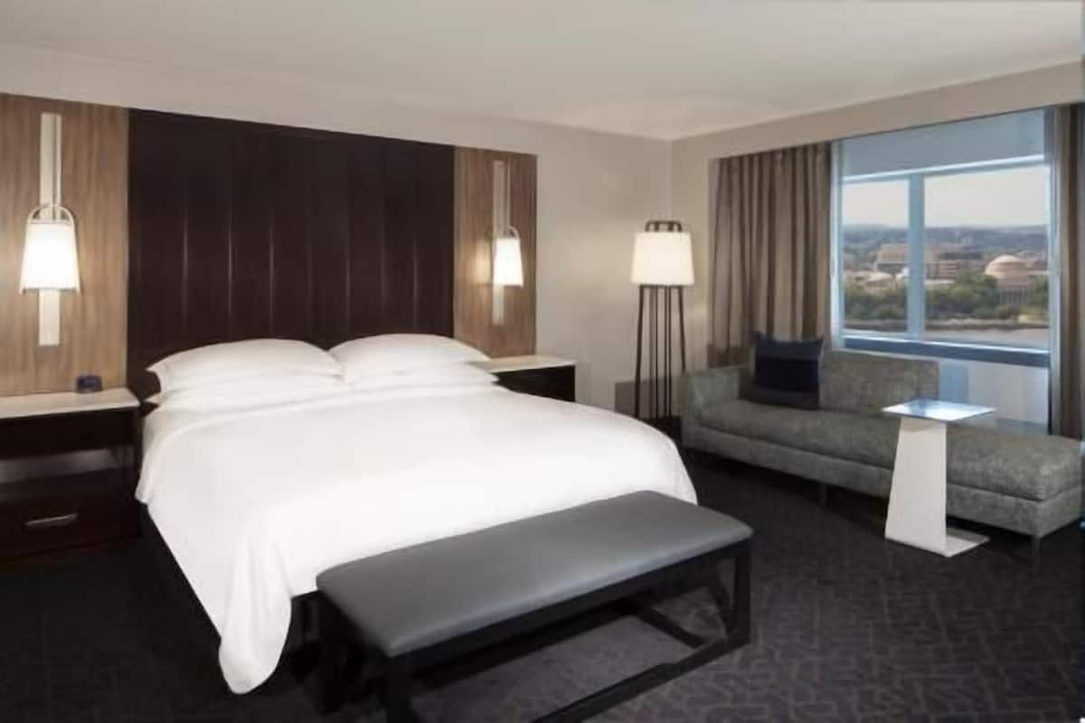 A hotel room with a bed, a bench, a sofa, a nightstand, a lamp, and a view of the city.