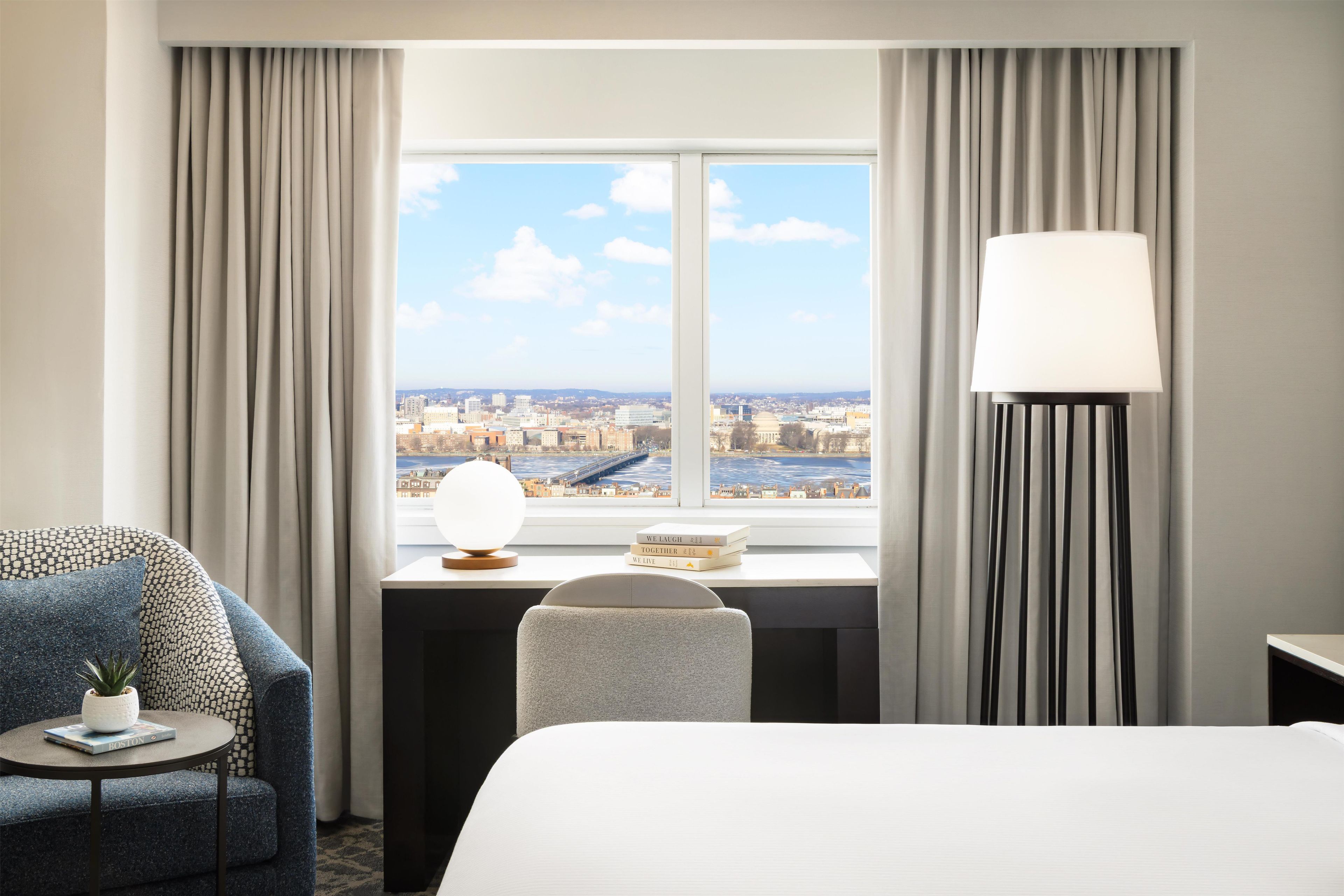 Ein Hotelzimmer mit Bett, Schreibtisch mit Lampe, Stuhl und Blick auf eine Stadtkulisse durch ein Fenster.