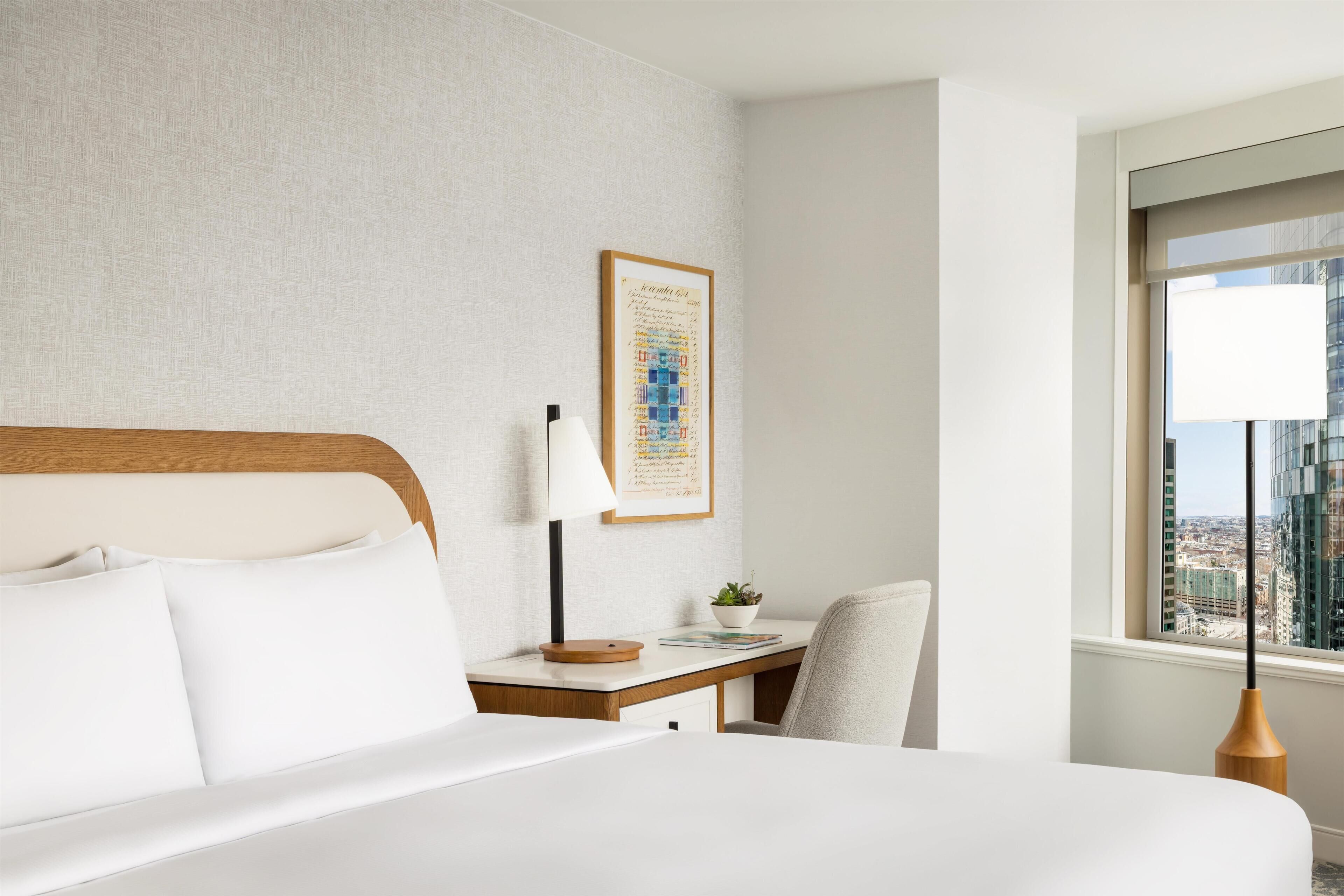 A hotel room with a bed, a desk, a chair, a lamp, a framed map, and a window with a city view.