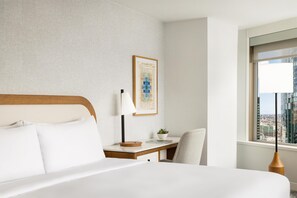 Et hotelværelse med seng, skrivebord, stol, lampe, et indrammet kort og et vindue med udsigt over byen.