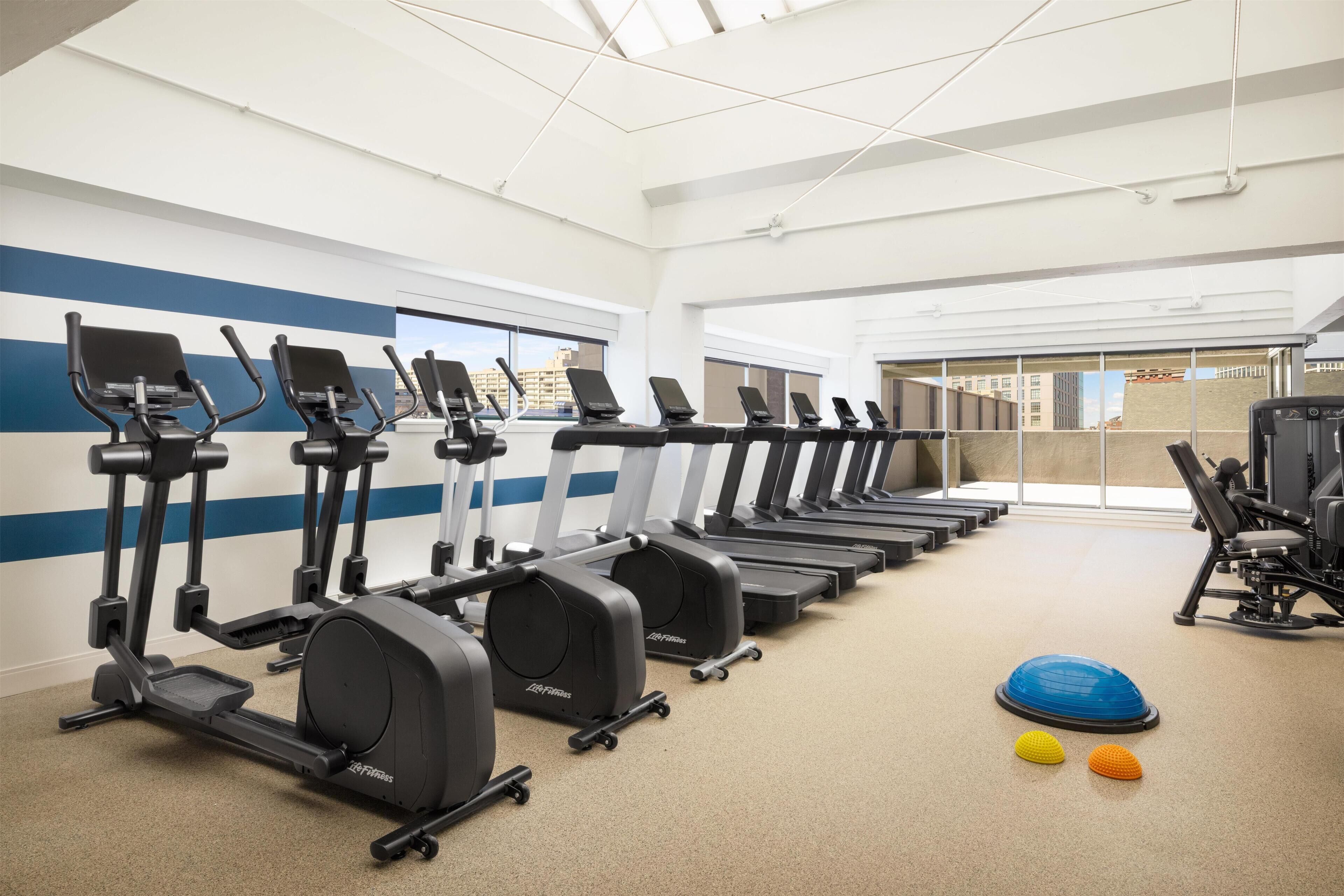 Un gimnasio con varias cintas de correr, pelotas de ejercicio y vistas de la ciudad a través de amplios ventanales.