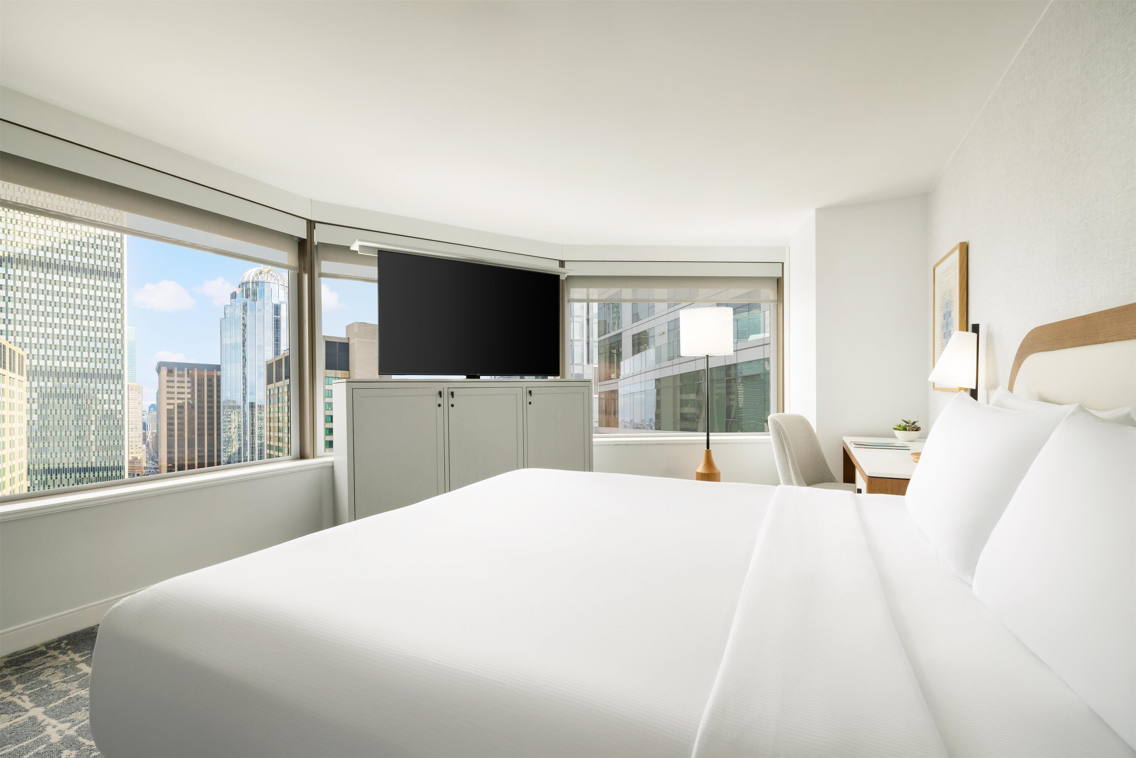 A hotel room with a large bed, a desk, a chair, a TV, and a window with a city view.