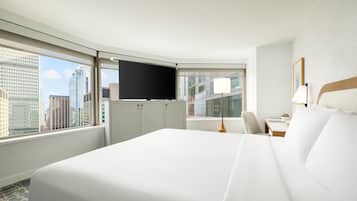 A hotel room with a large bed, a desk, a chair, a TV, and a window with a city view.