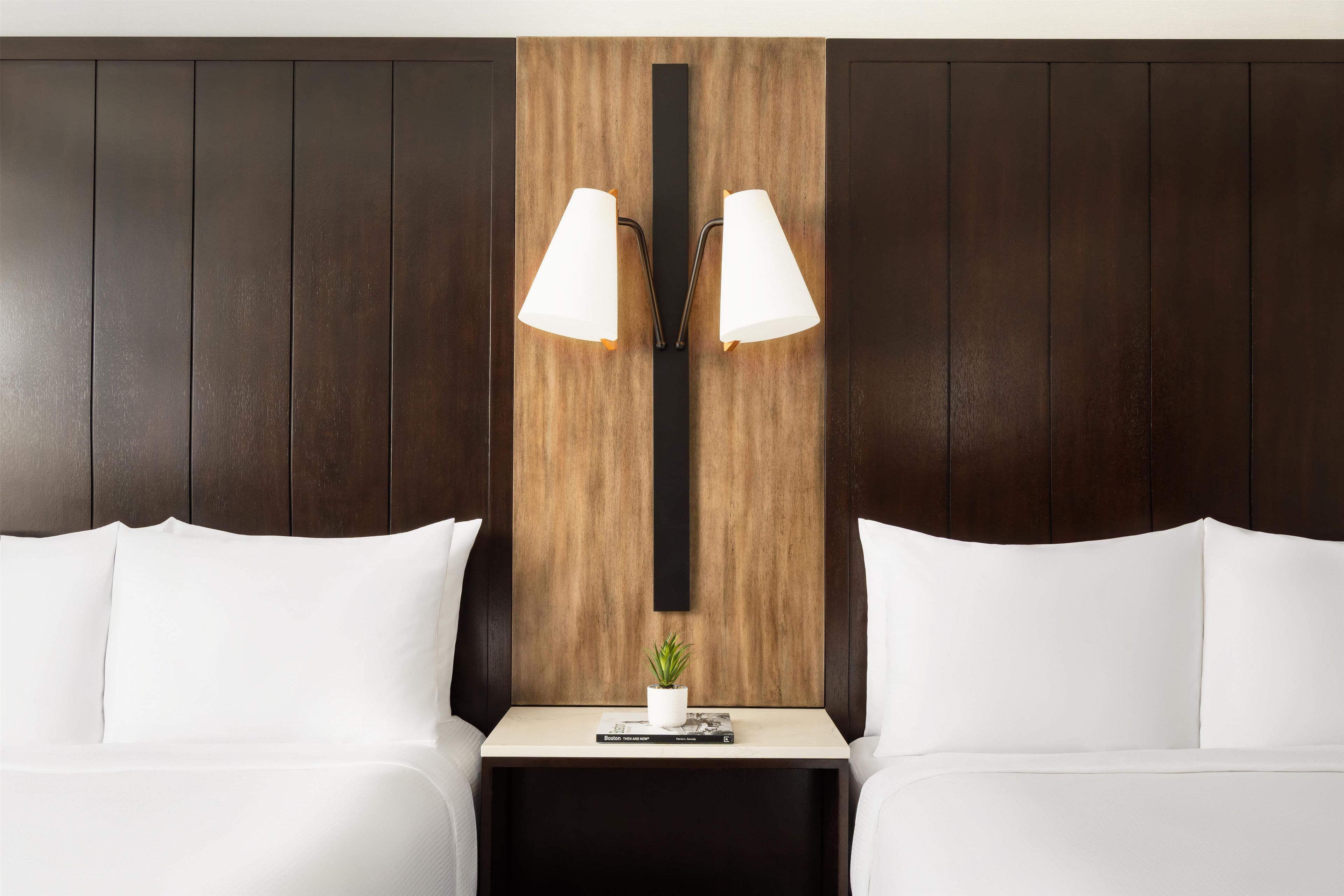 Ein Hotelzimmer mit dunkelholzigem Kopfteil, zwei weißen Kissen, einem kleinen Nachttisch mit Buch und Pflanze sowie zwei Wandlampen.