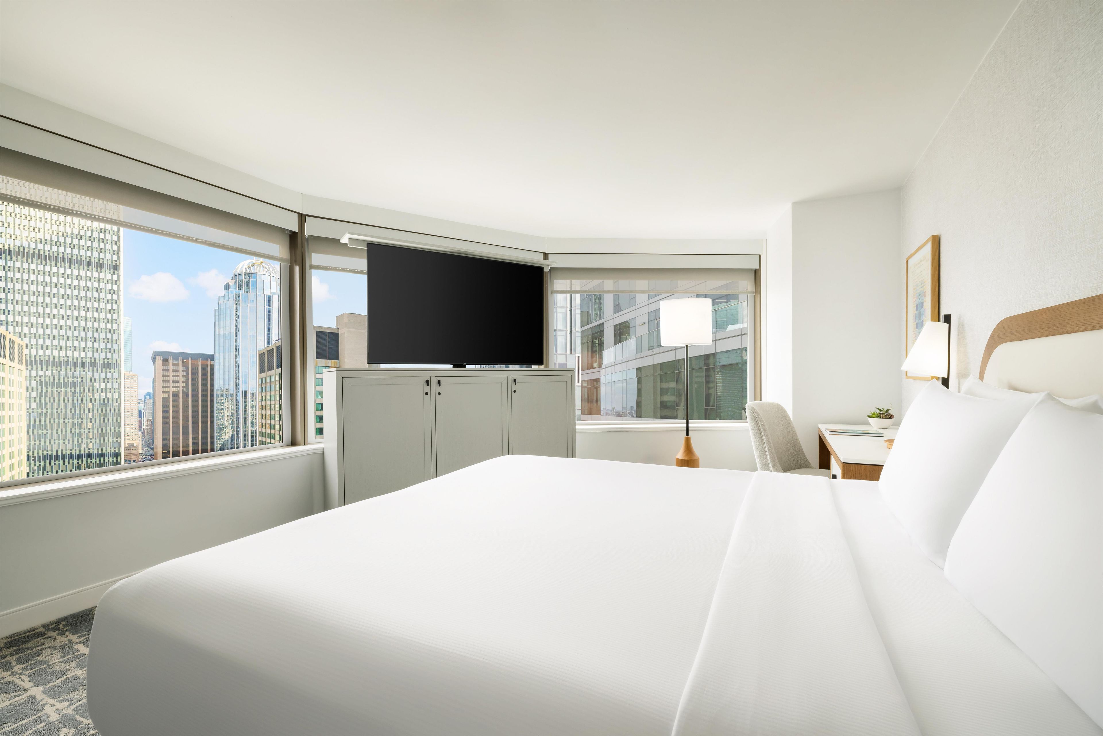 A hotel room with a large bed, a desk, a chair, a TV, and a window with a city view.