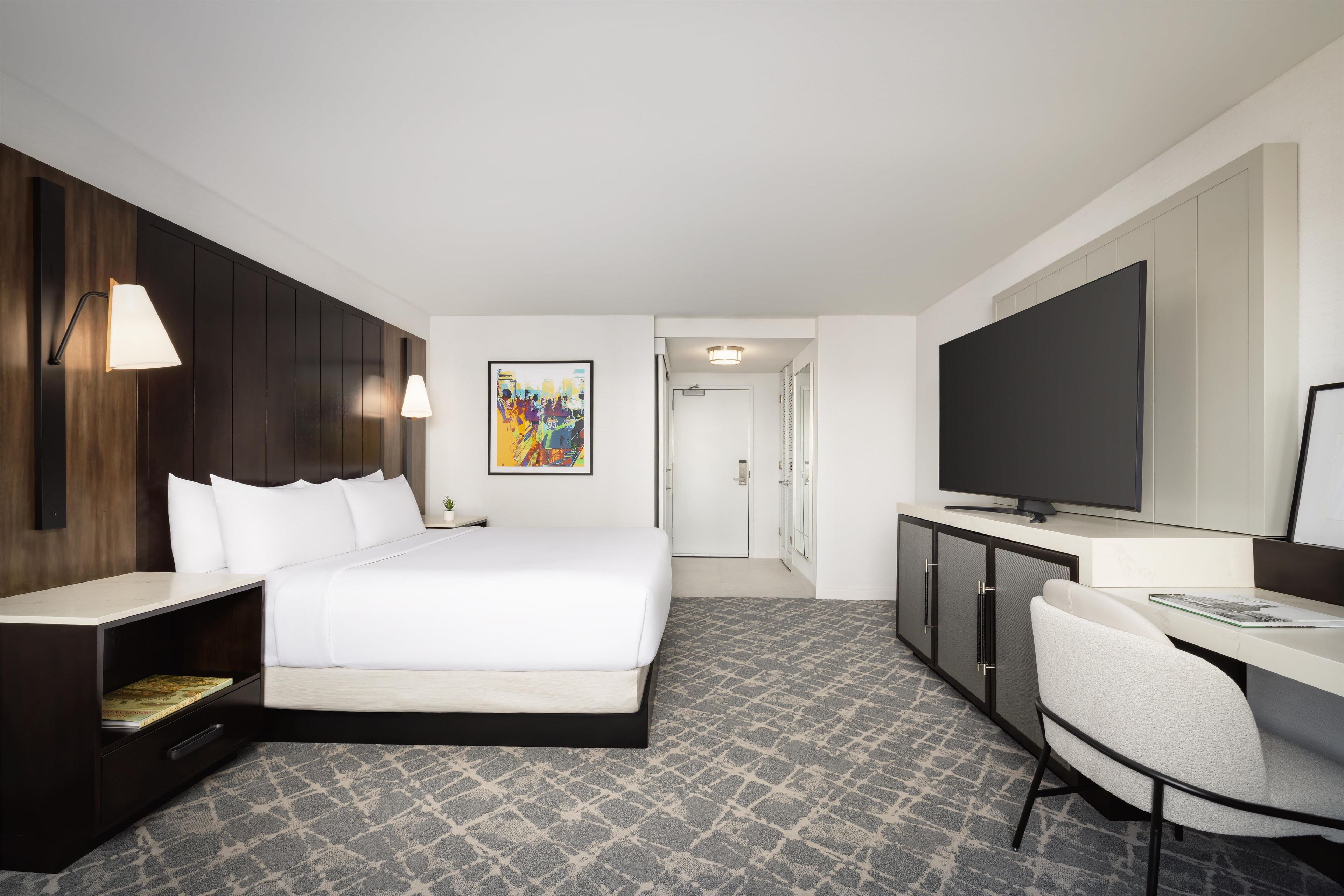 A hotel room with a bed, bedside table, TV, desk, chair, and artwork on the wall.