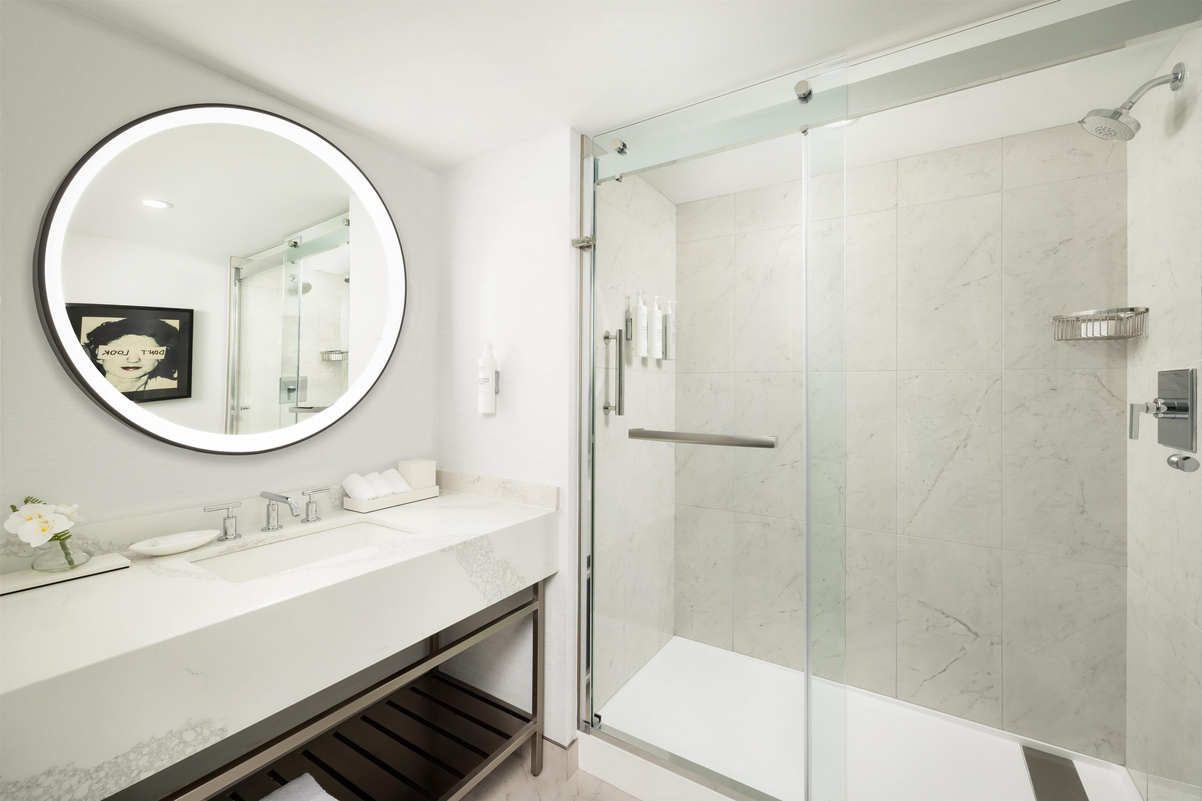 A modern bathroom with a glass-enclosed shower, a round mirror, and a marble countertop.