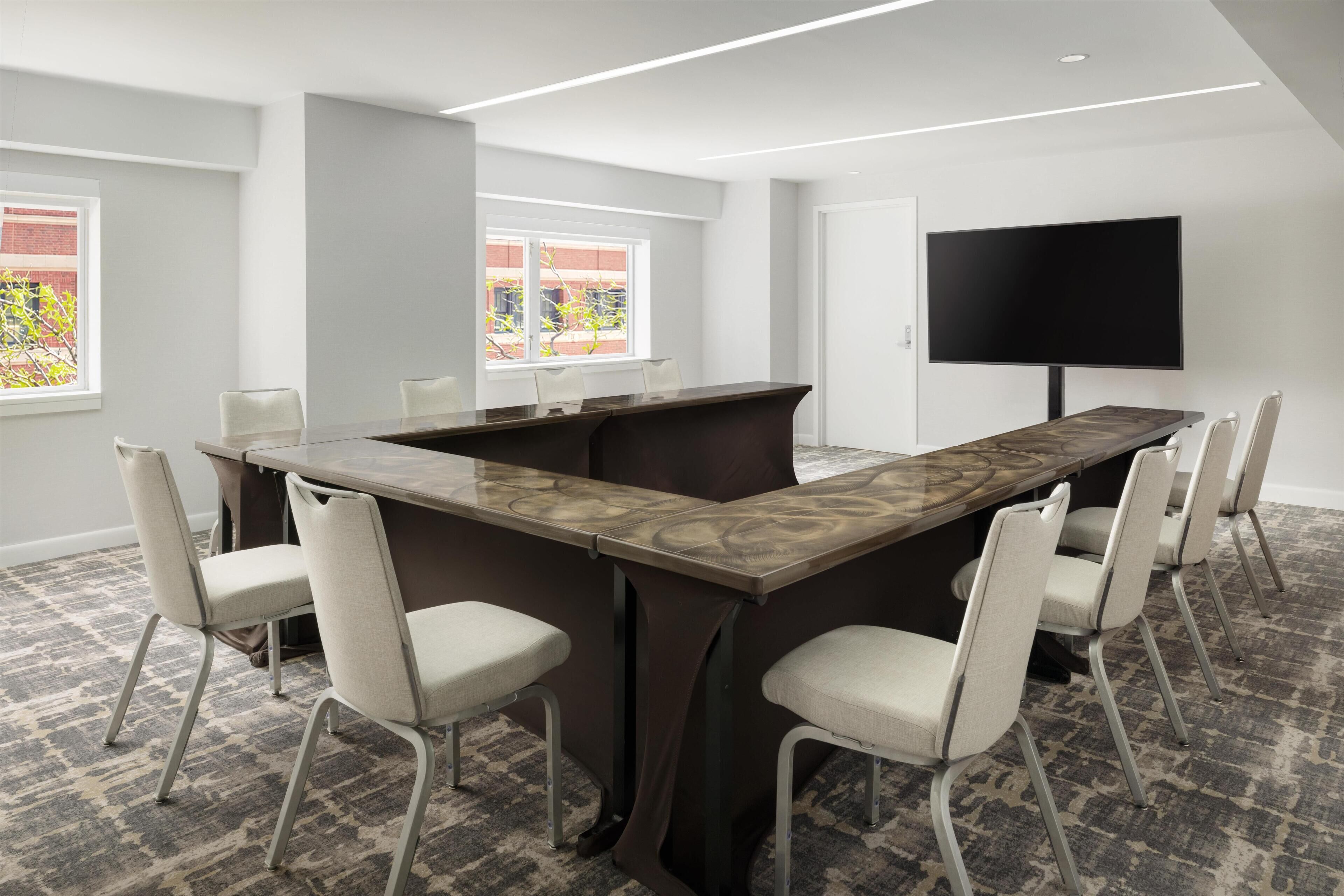 A conference room with a long table, six chairs, a large flat-screen TV, and windows with a view of greenery.