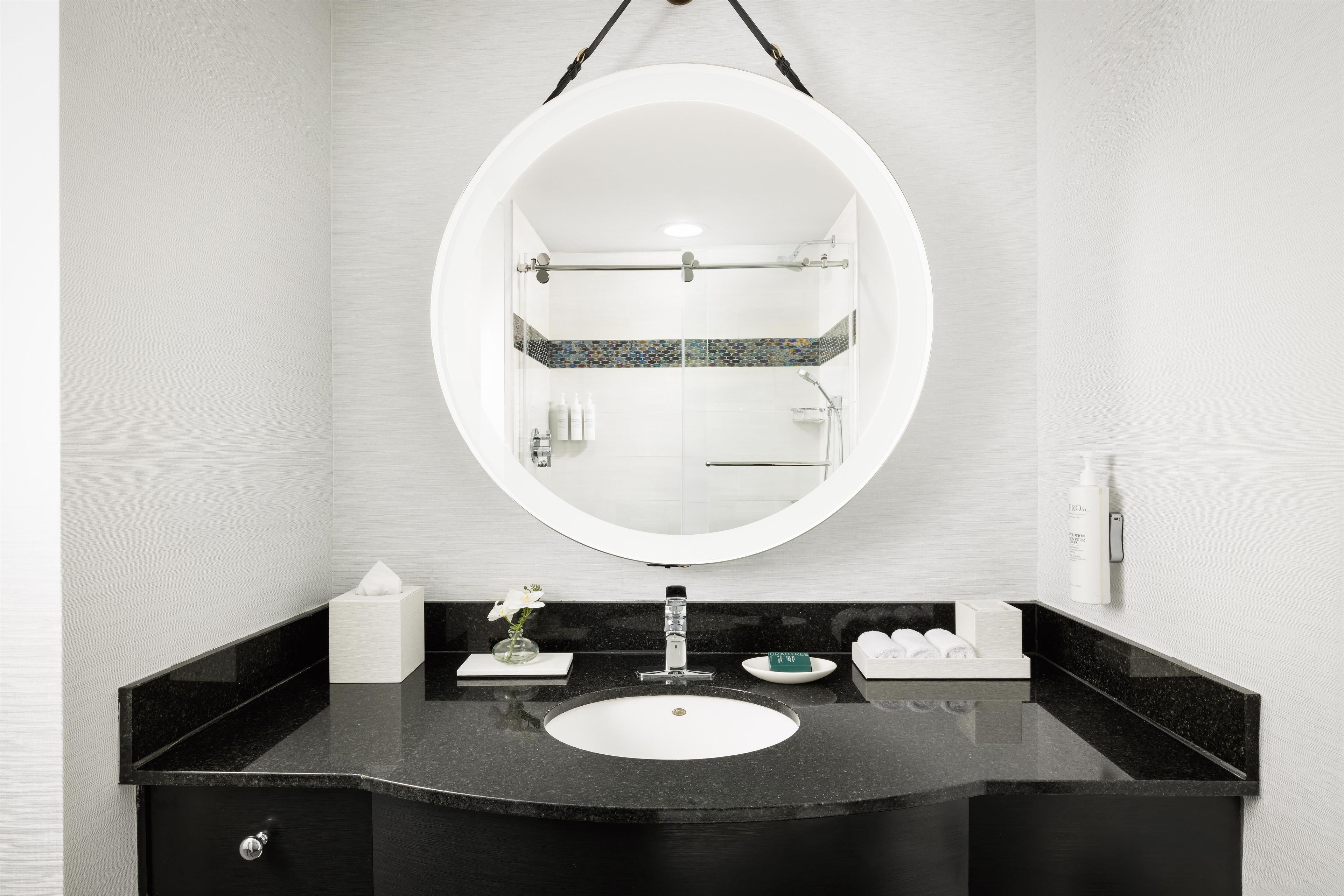 A bathroom with a round mirror, a sink, a towel dispenser, and a soap dispenser.