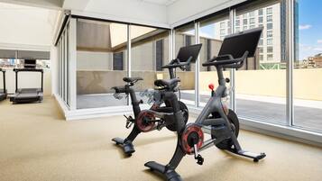 A gym with stationary bikes, a treadmill, and large windows offering a city view.
