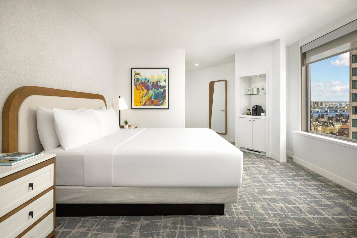 A hotel room with a bed, a nightstand, a mirror, a painting, and a view of the city.
