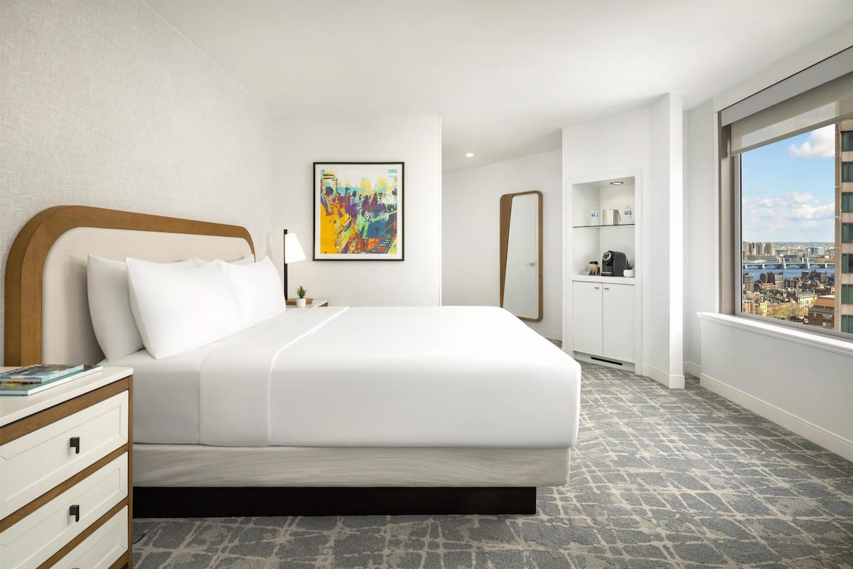 A hotel room with a bed, a nightstand, a mirror, a painting, and a view of the city.