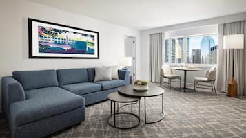 A modern hotel room with a sectional sofa, a small round table, a dining area with chairs, a large window with a city view, and a framed artwork on the wall.