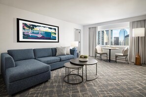 Una habitación de hotel moderna con un sofá modular, una pequeña mesa redonda, un rincón de comedor con sillas, un ventanal con vista a la ciudad y una obra de arte enmarcada en la pared.