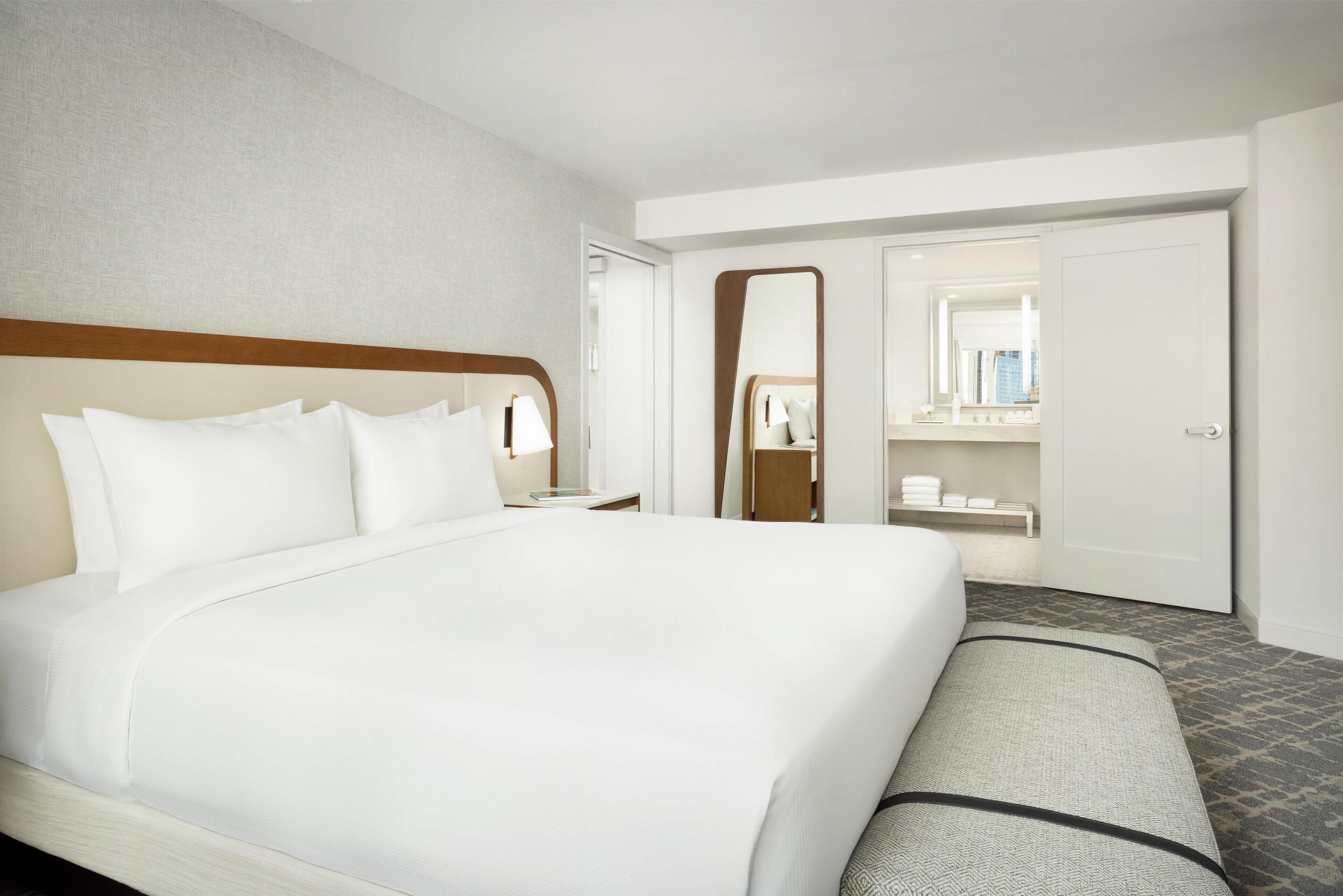 Une chambre d’hôtel avec un grand lit, une table de chevet, un miroir, une armoire et une salle de bain visible à travers une porte ouverte.