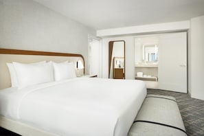 Une chambre d’hôtel avec un grand lit, une table de chevet, un miroir, une armoire et une salle de bain visible à travers une porte ouverte.