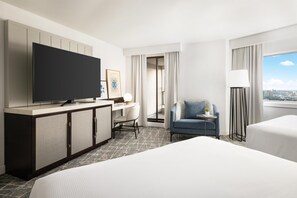 A hotel room with a bed, a desk with a chair, a television, a small table with a lamp, and a view of the city through the window.