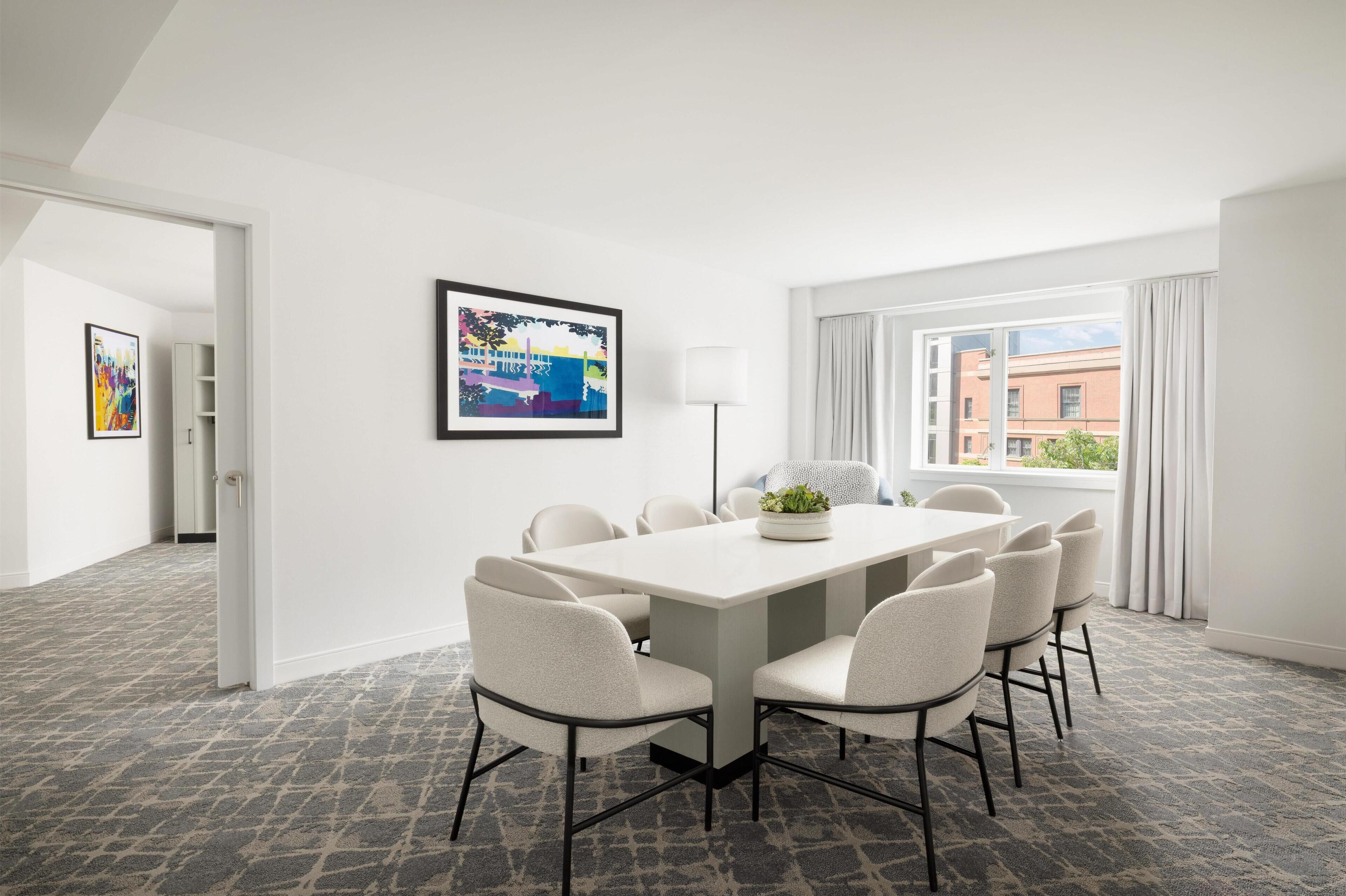 A modern hotel room with a large dining table, six chairs, a floor lamp, a framed picture, and a view of buildings through the window.
