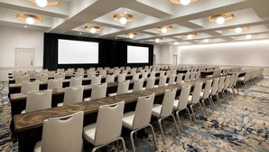 Un salón de conferencias con filas de mesas y sillas, una pantalla grande y luces en el techo.