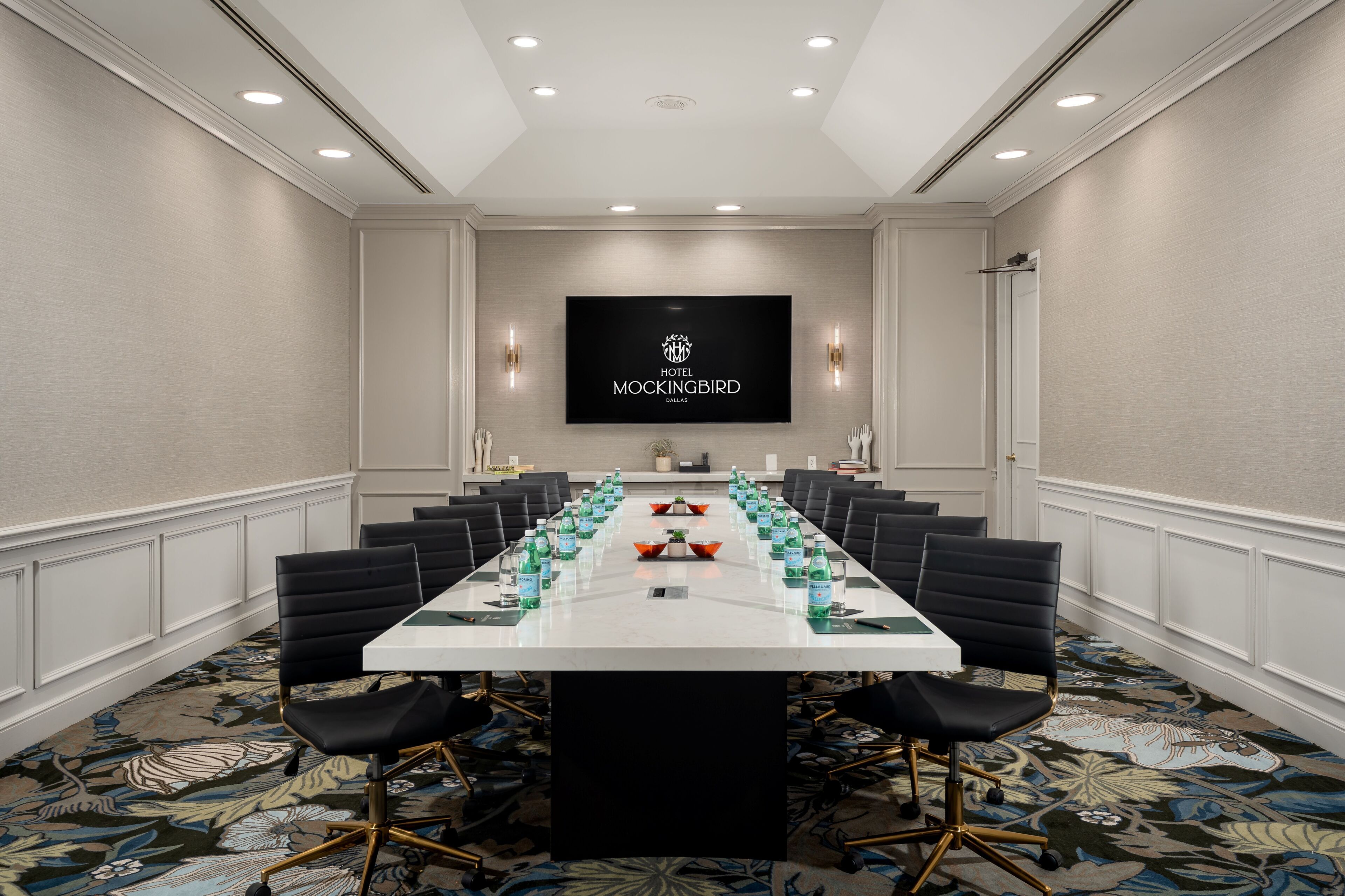 A conference room with a long table, black chairs, and a flat-screen TV displaying 'Hotel Mockingbird Dallas.'