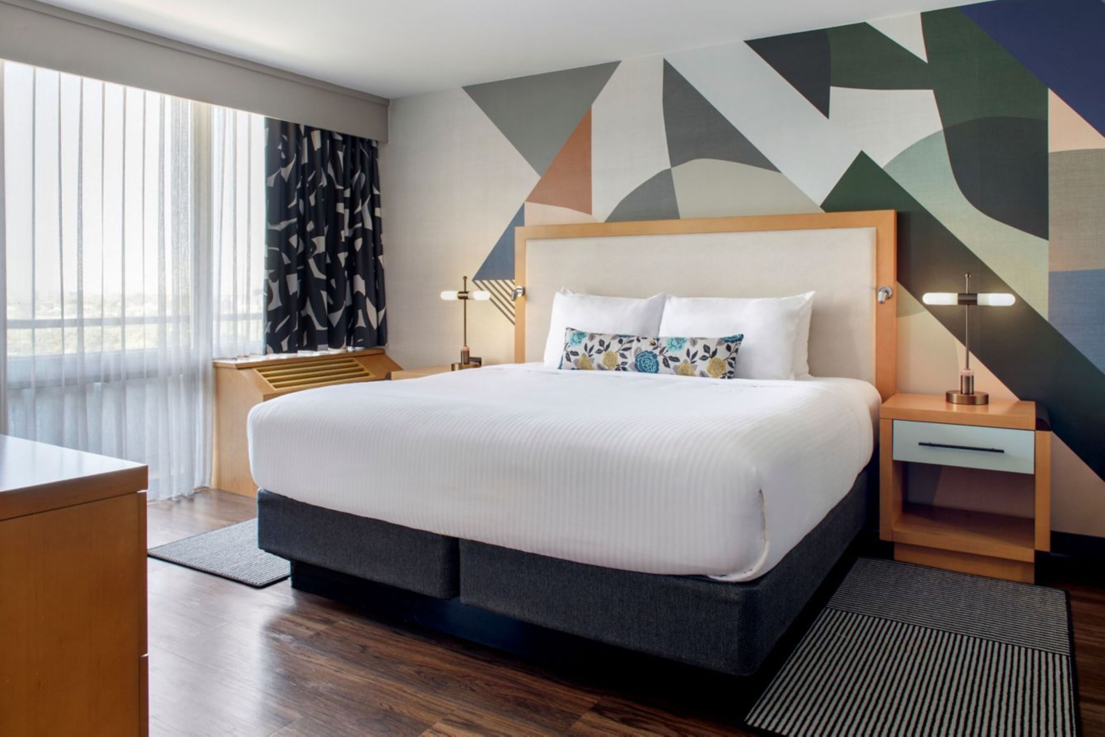 A hotel room with a large bed, a wooden nightstand, a desk, and a view through the window.