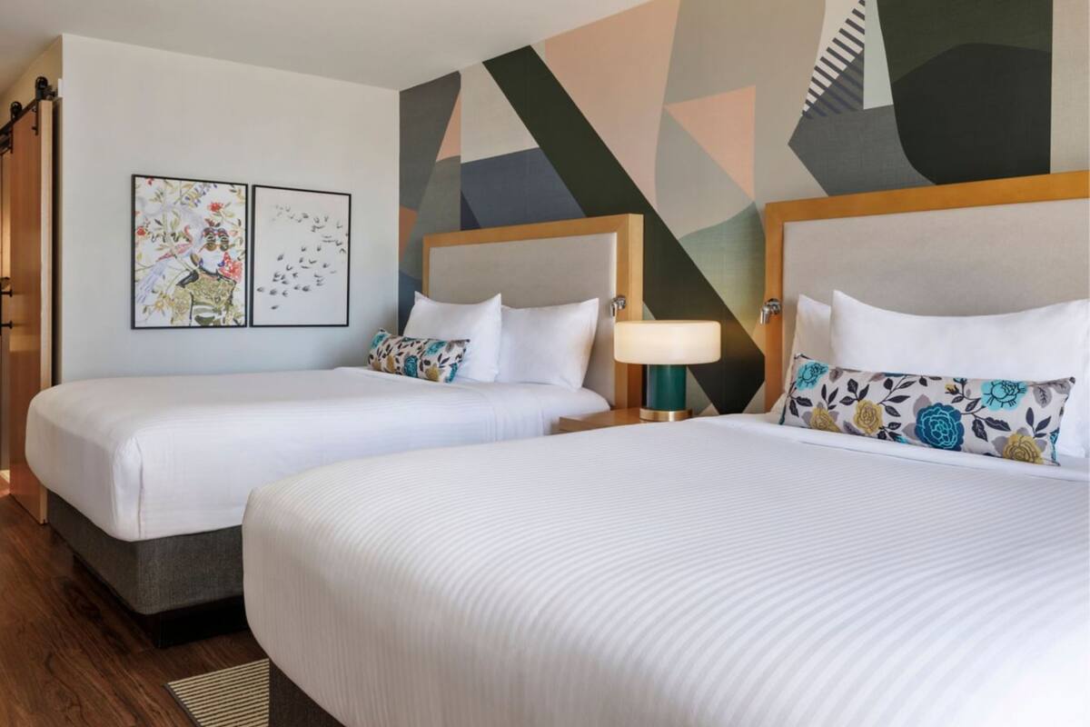 A hotel room with two beds, a nightstand with a lamp, and abstract wall art.