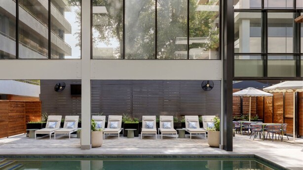 Zona de piscina con sillones de descanso y sombrillas, rodeada por un edificio moderno con grandes ventanales.