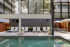 A pool area with lounge chairs and umbrellas, surrounded by a modern building with large windows.