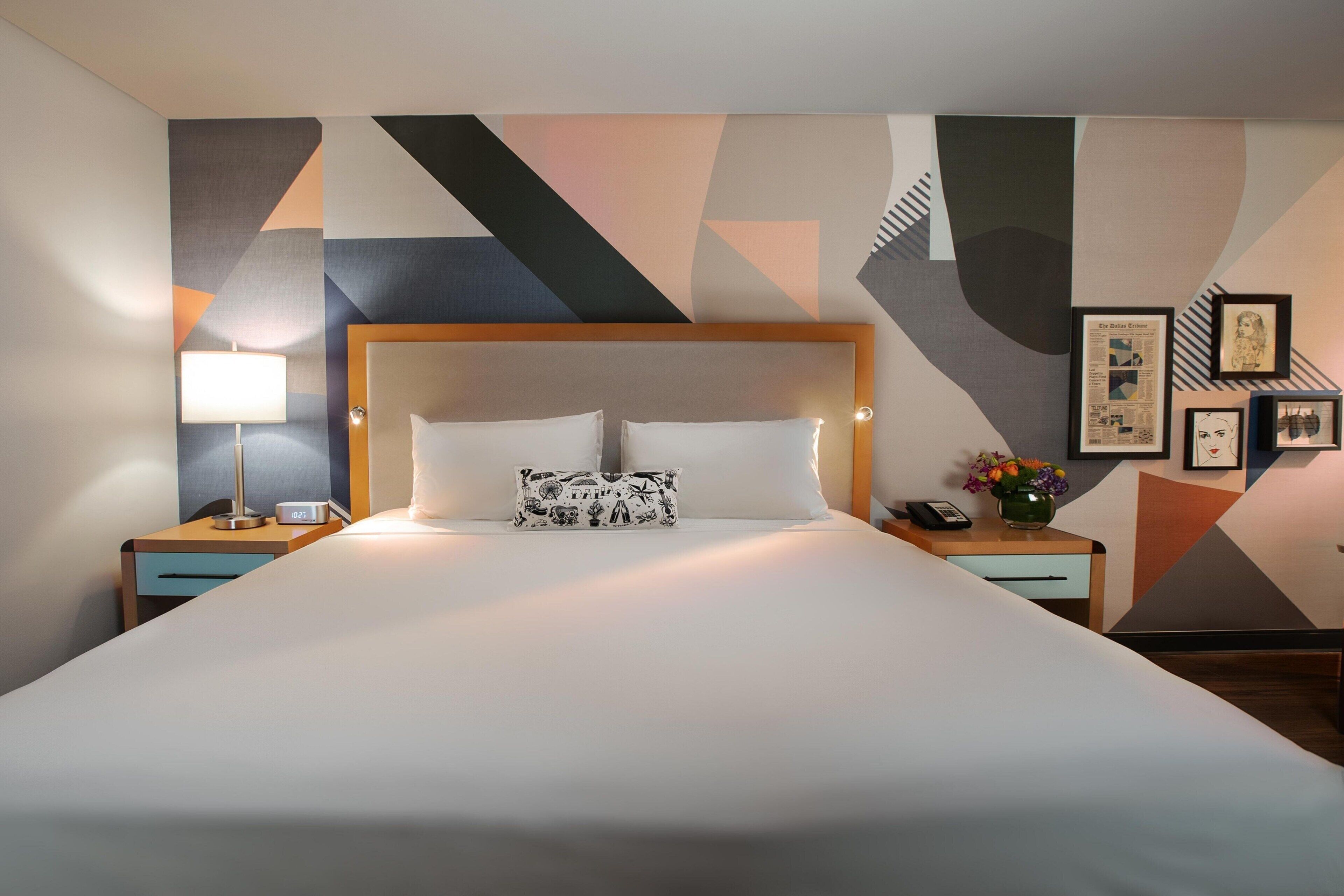 A hotel room with a large bed, a nightstand with a lamp, a phone, and a framed picture on the wall.