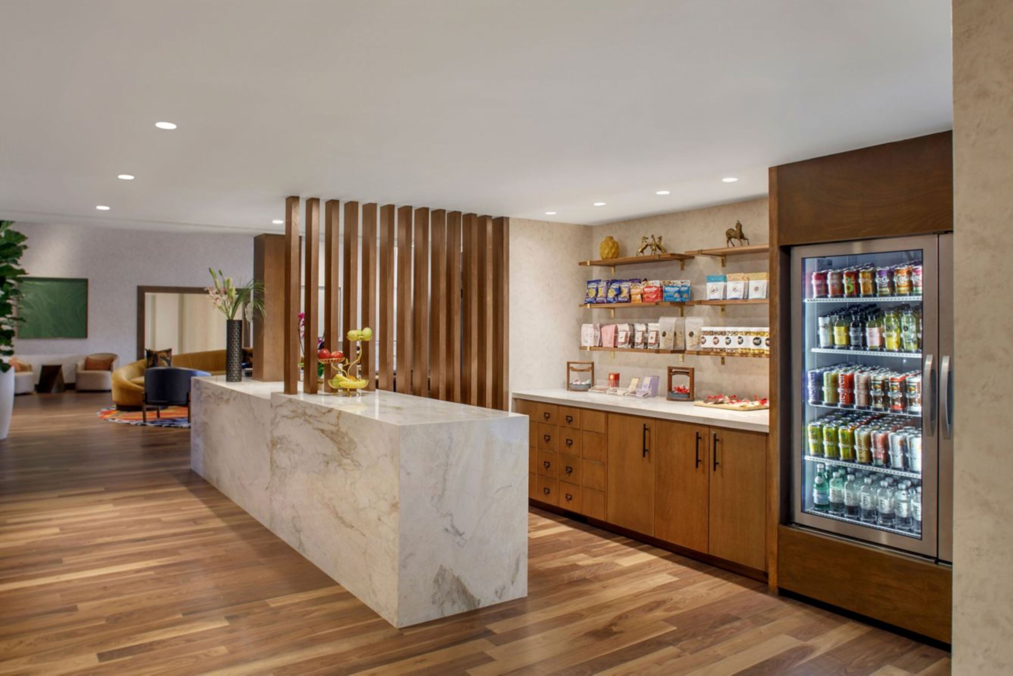 A hotel lobby with a marble reception desk, wooden cabinets, a refrigerator stocked with beverages, and a seating area with a sofa and chairs.