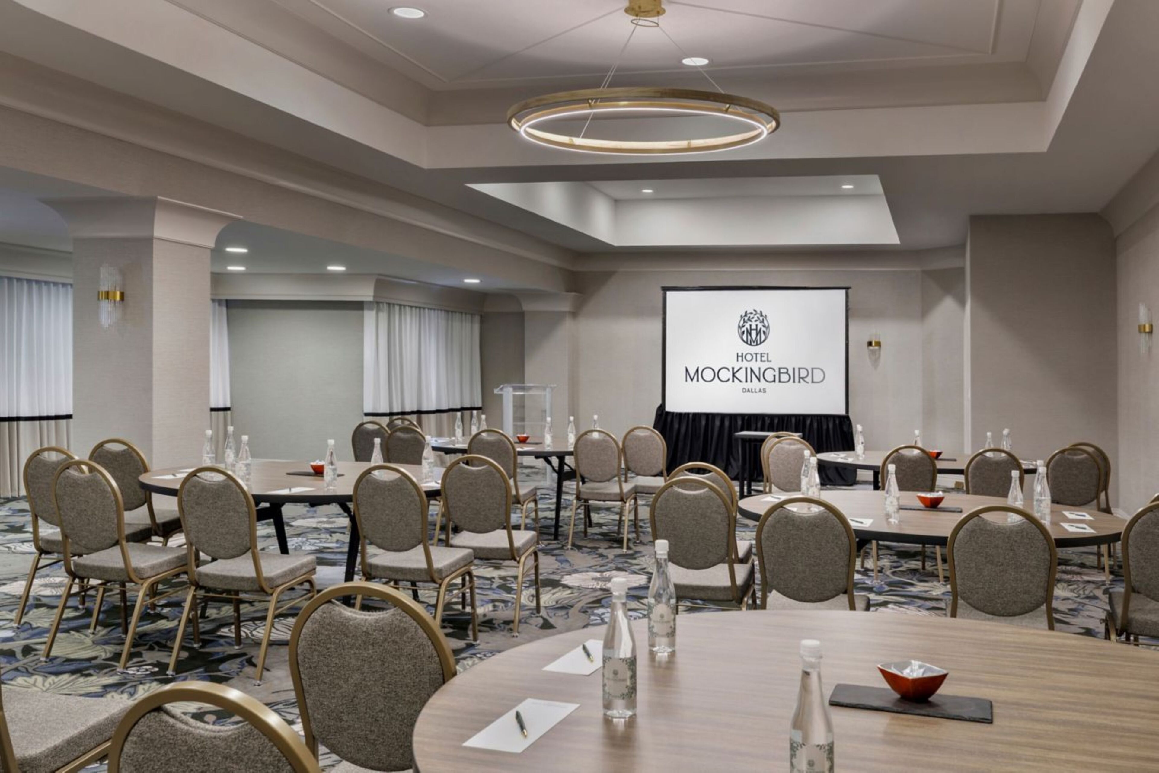 Un salón de conferencias con mesas redondas, sillas y una pantalla de proyección mostrando 'HOTEL MOCKINGBIRD DALLAS'.