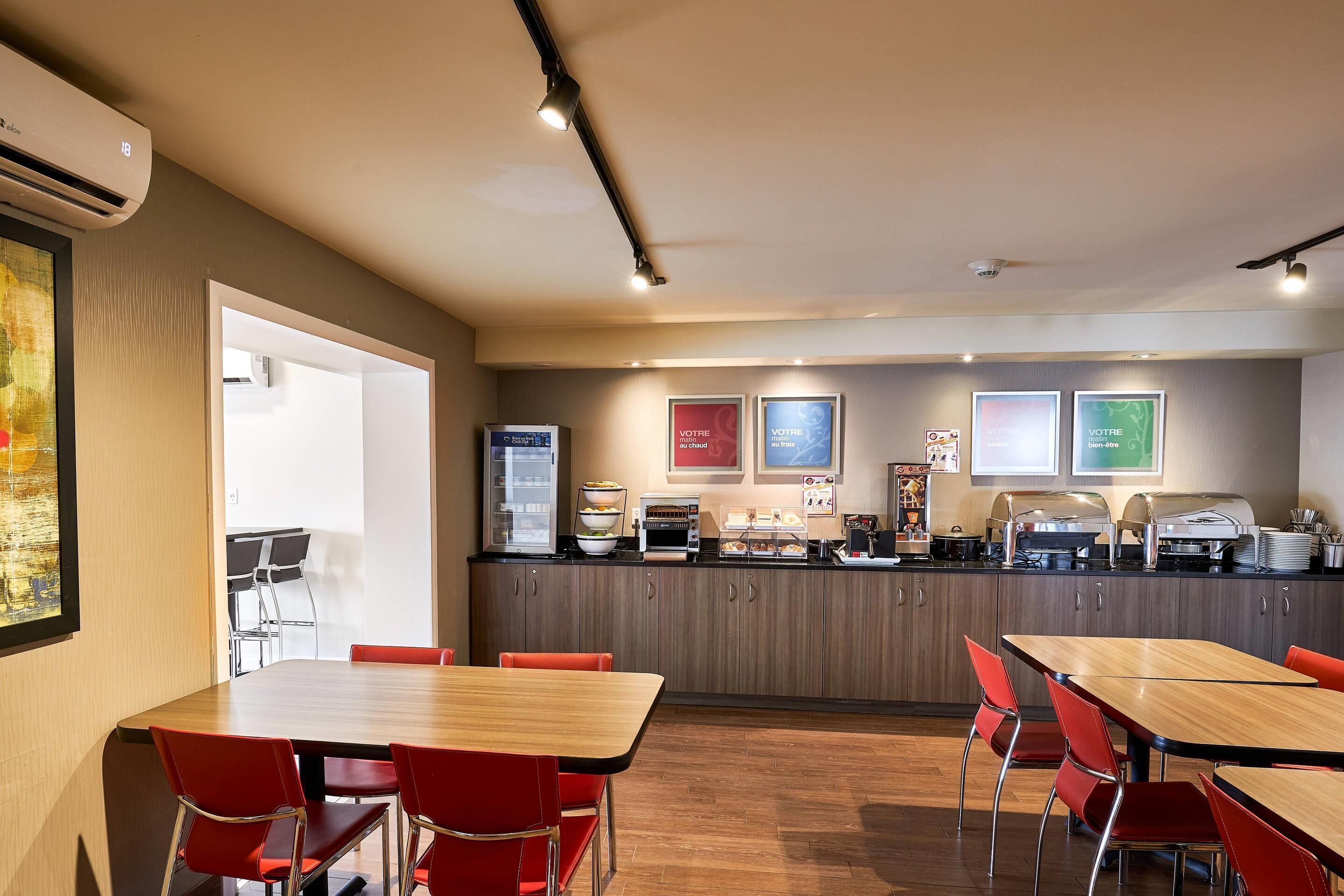 A modern dining area with wooden tables and red chairs, a kitchen counter with appliances, and framed pictures on the wall.