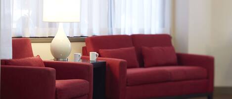 Una habitación con un sofá rojo, un sillón rojo más pequeño, una mesa con una lámpara y una ventana con cortinas traslúcidas.