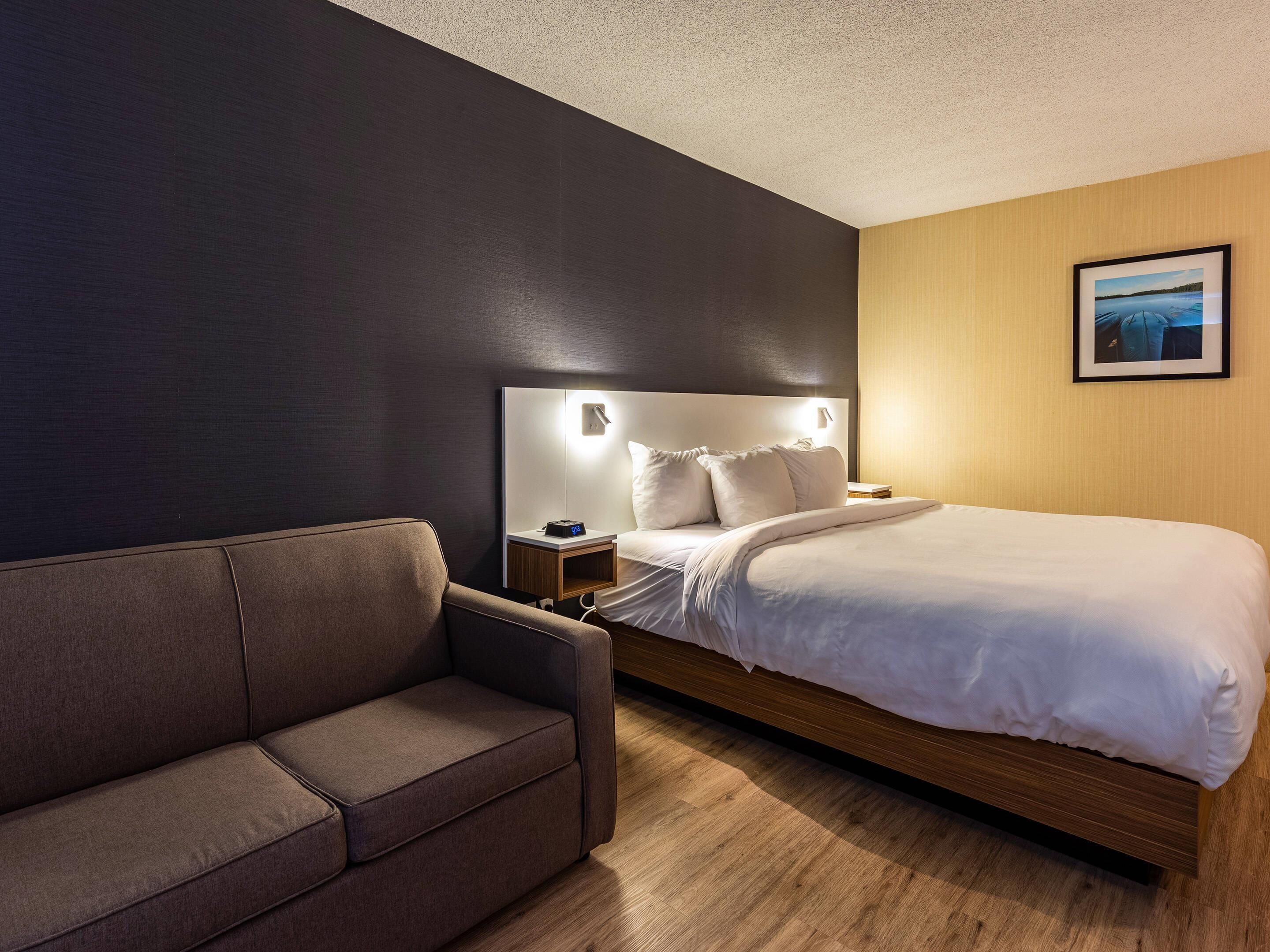A modern hotel room with a bed, a nightstand, a wall-mounted lamp, a sofa, and a framed picture on the wall.