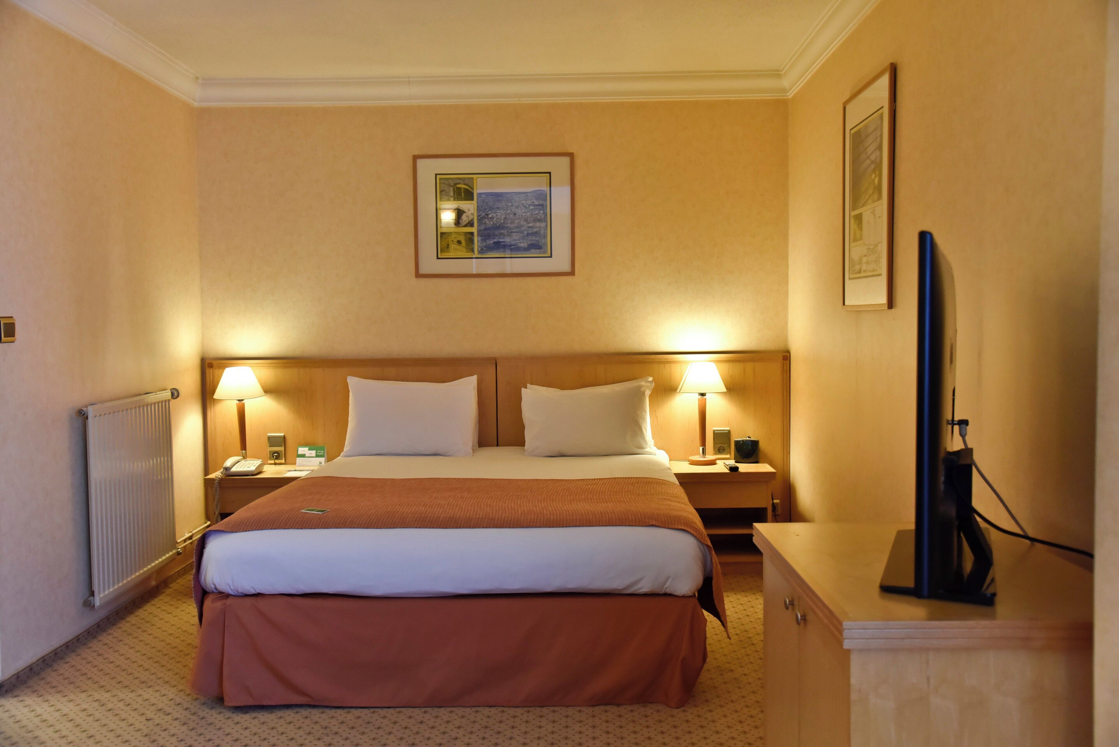 A hotel room with a large bed, two bedside lamps, a flat-screen TV, and a framed picture on the wall.