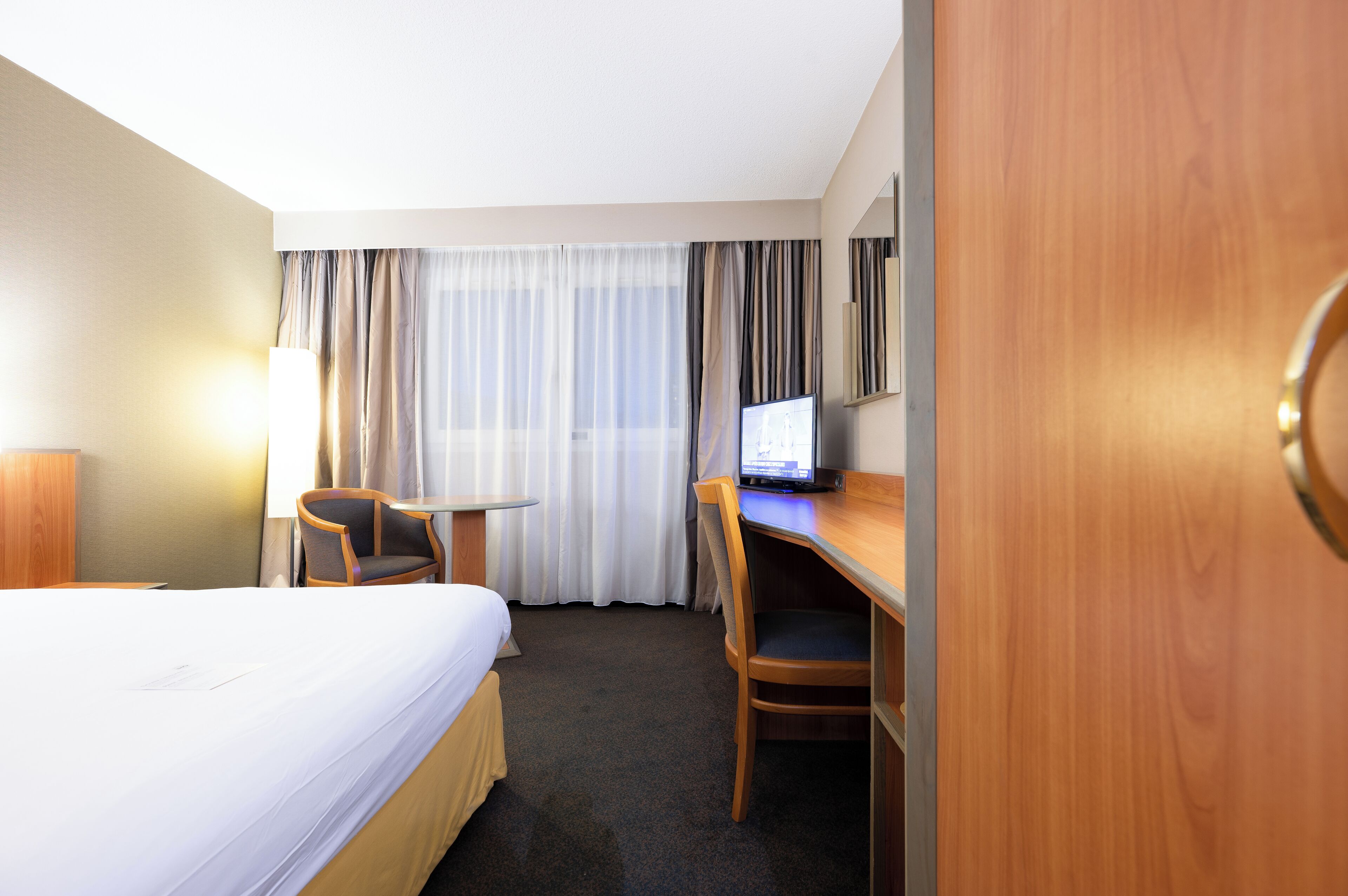 Una habitación de hotel con cama, escritorio, silla, televisor y ventana con cortinas.