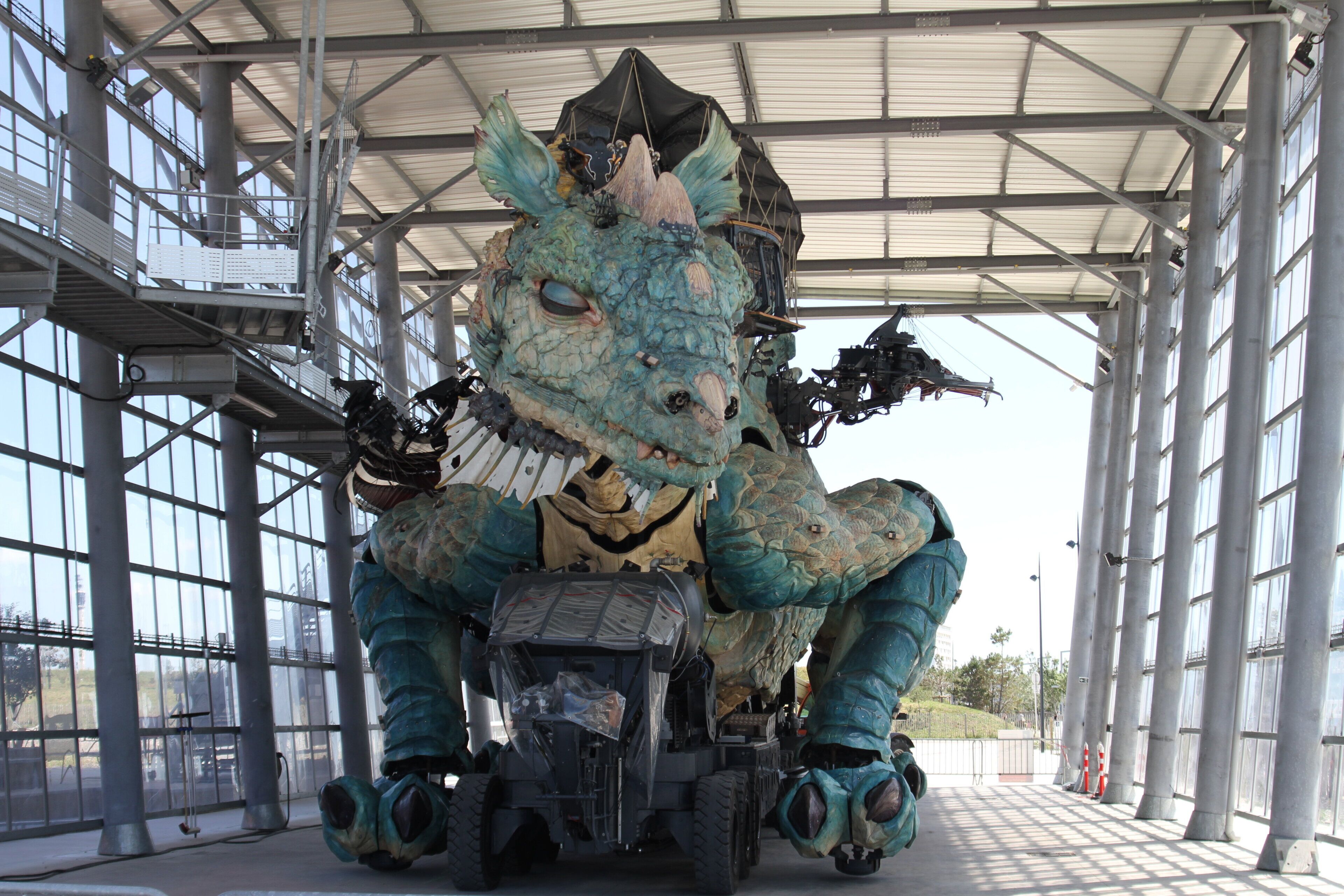 Un grand véhicule immobile, arborant un design rappelant un dragon, est stationné sous une structure abritée dotée de grandes fenêtres.