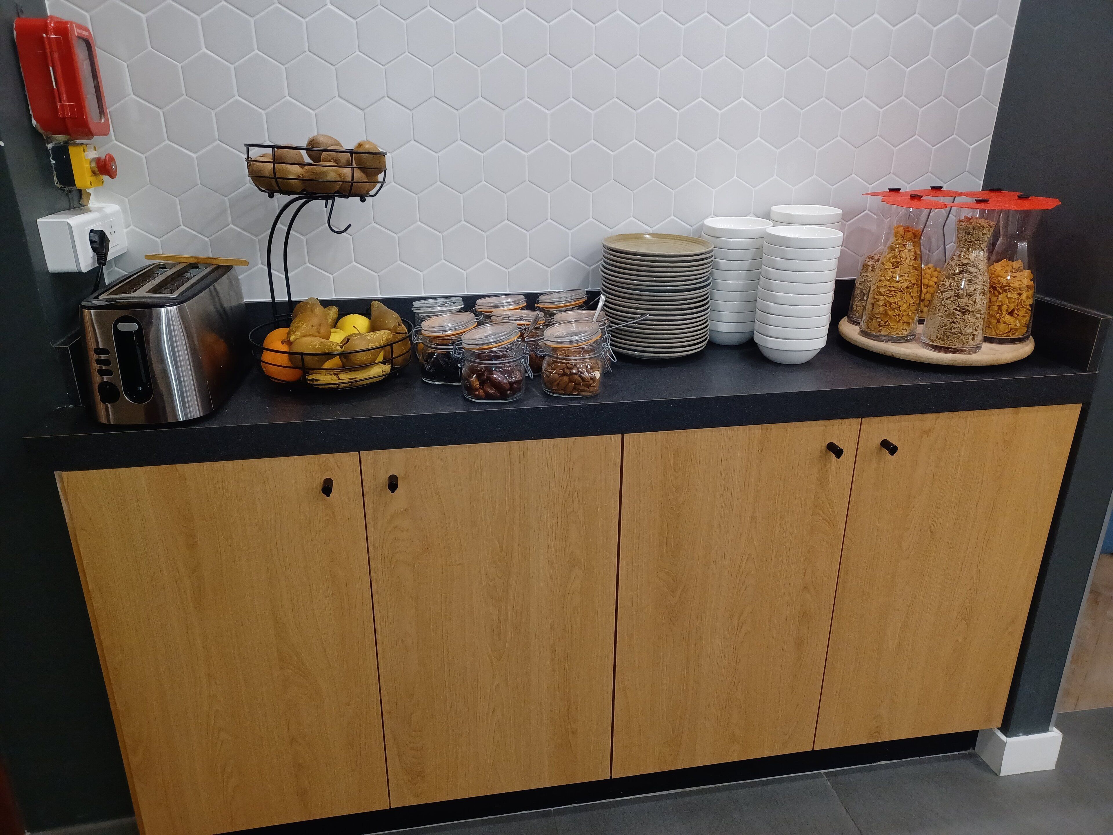 A kitchen counter with a toaster, a fruit basket, jars of nuts, and stacks of plates.