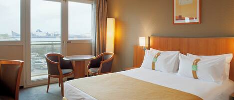 A hotel room with a bed, two chairs, a small table, a lamp, and a window with a view of the city.