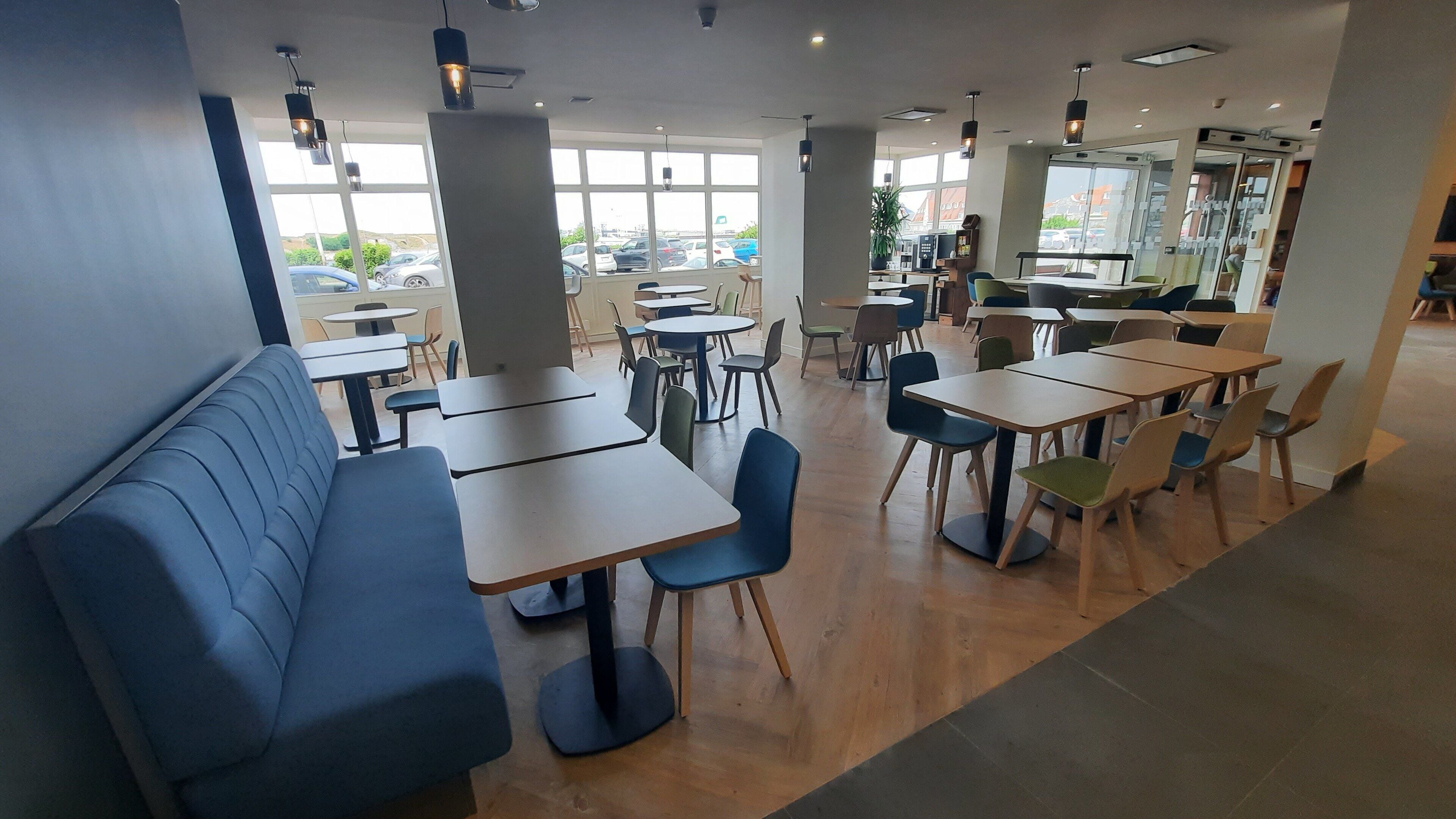 Um restaurante espaçoso com mesas e cadeiras altas, área de bancos azuis e grandes janelas com vista para o estacionamento.