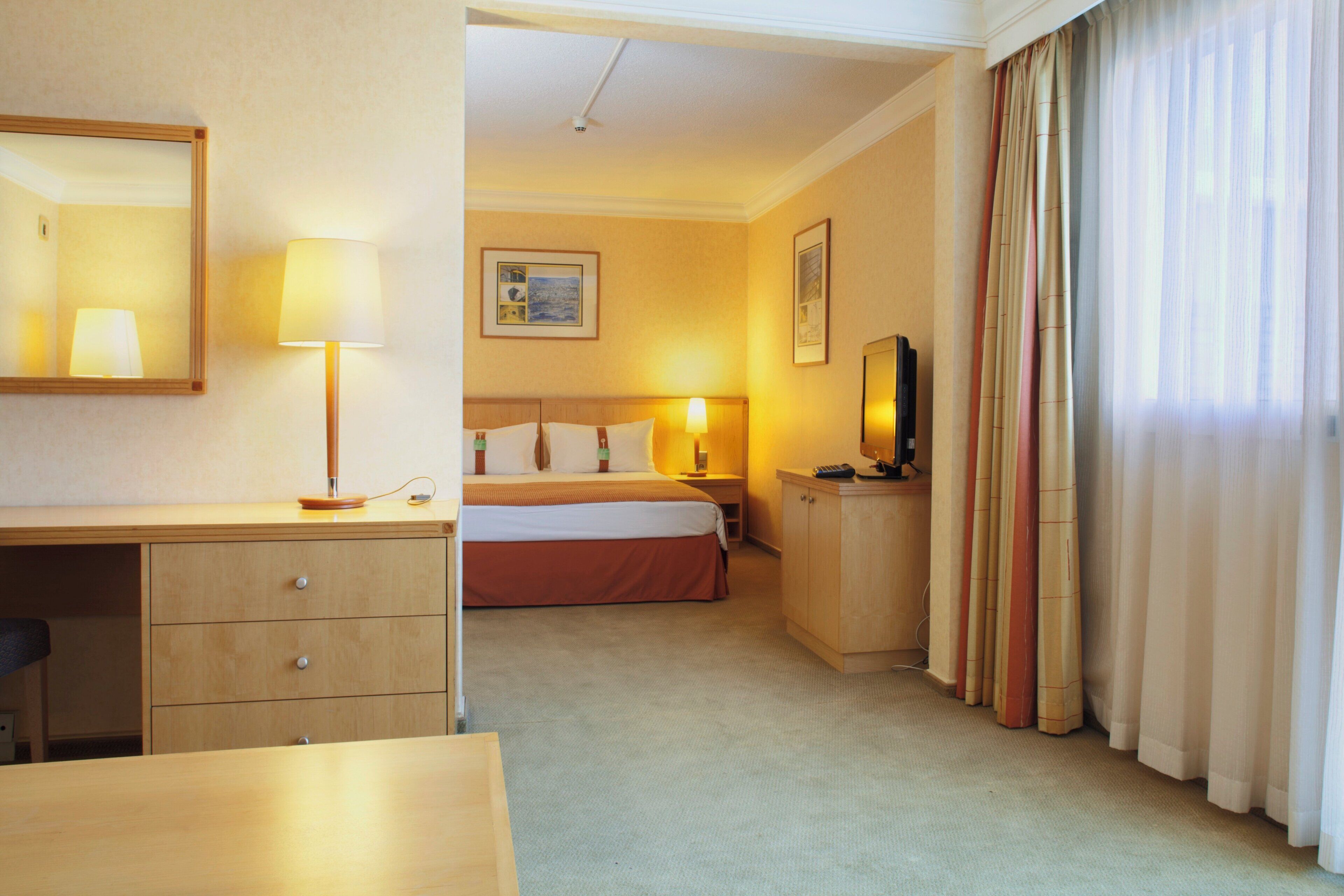 Habitación de hotel con cama, escritorio con lámpara, mesita de noche con lámpara, espejo, armario y ventana con cortinas.