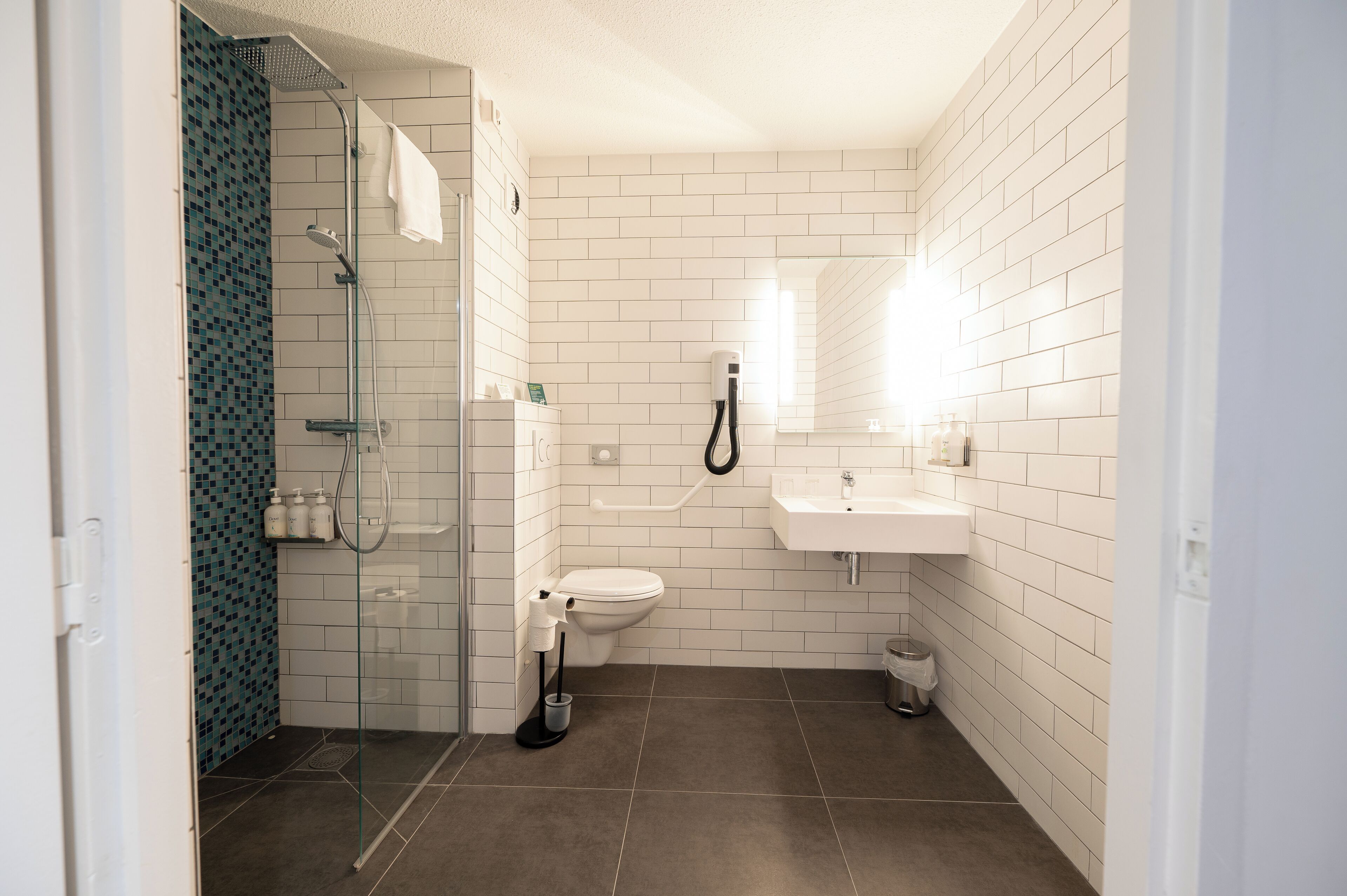 Un baño moderno con cabina de ducha de vidrio, paredes cubiertas de azulejos blancos, un inodoro y un lavamanos.
