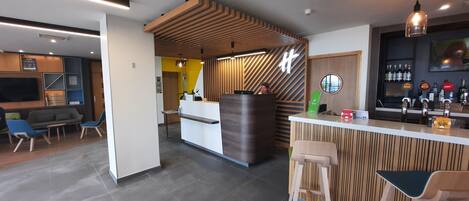 A modern bar area with a wooden reception desk, bar stools, and a seating area with a sofa and chairs.